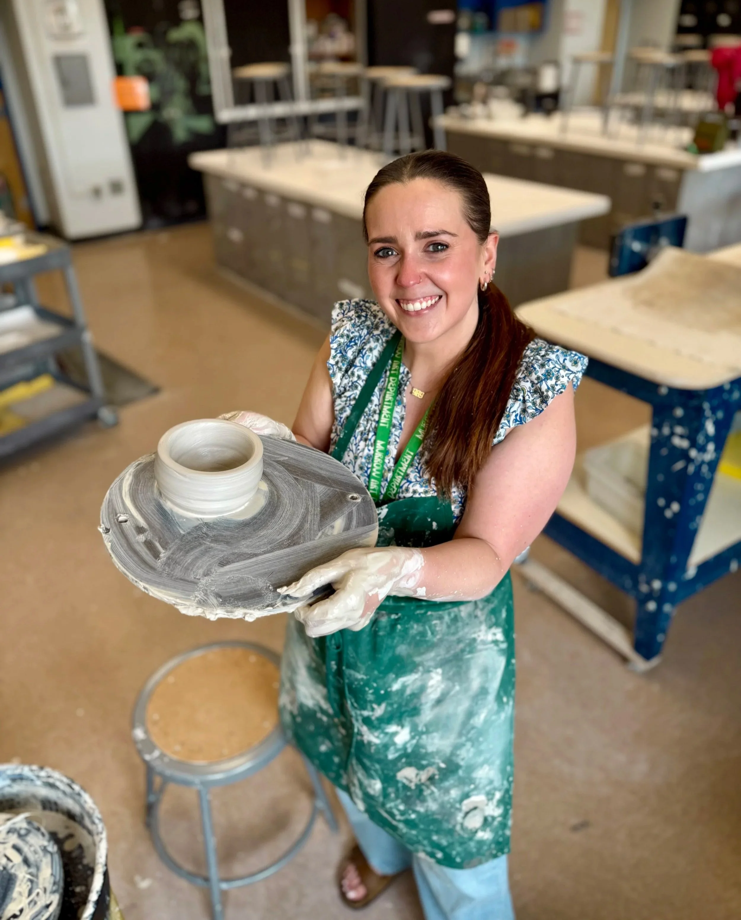 A woman with long brown hair, wearing a green apron and a lanyard, is holding a pottery wheel with a ceramic piece in progress in a pottery studio.