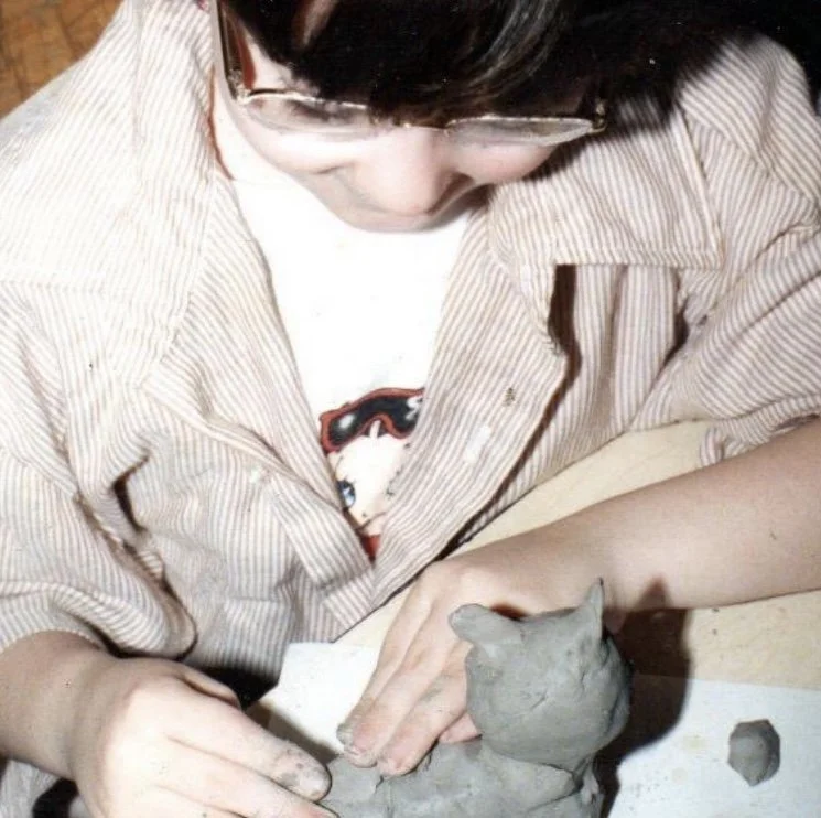 A young person wearing glasses and a striped shirt working with clay, shaping a small clay figure of a cat on a table.