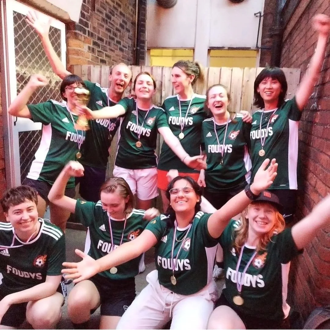 A group of ten young people celebrating after a sports event, wearing green jerseys with the word 'FOUDYS' and medals around their necks, posing cheerfully in an outdoor brick alleyway.
