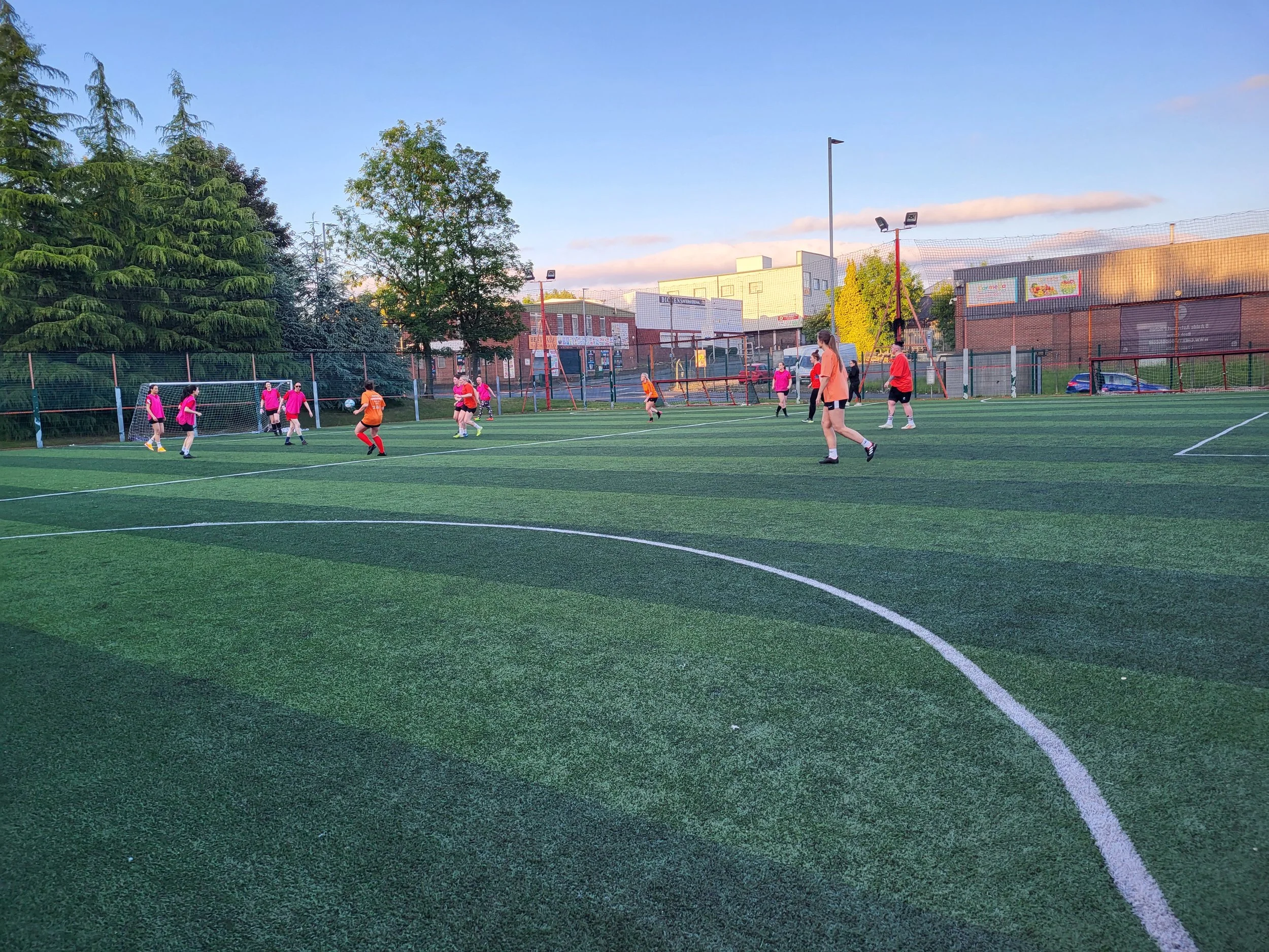 People playing soccer on an outdoor field during the evening, with trees and buildings in the background.