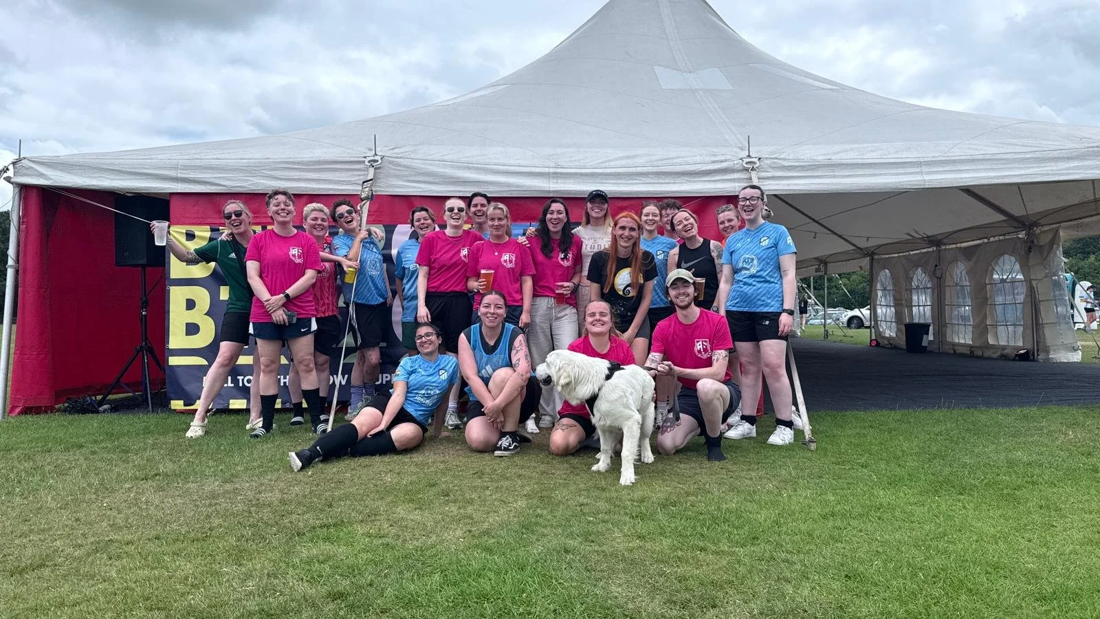 Group of players in colorful sports attire posing for a photo at outdoor event under a large white tent, with a dog in front.