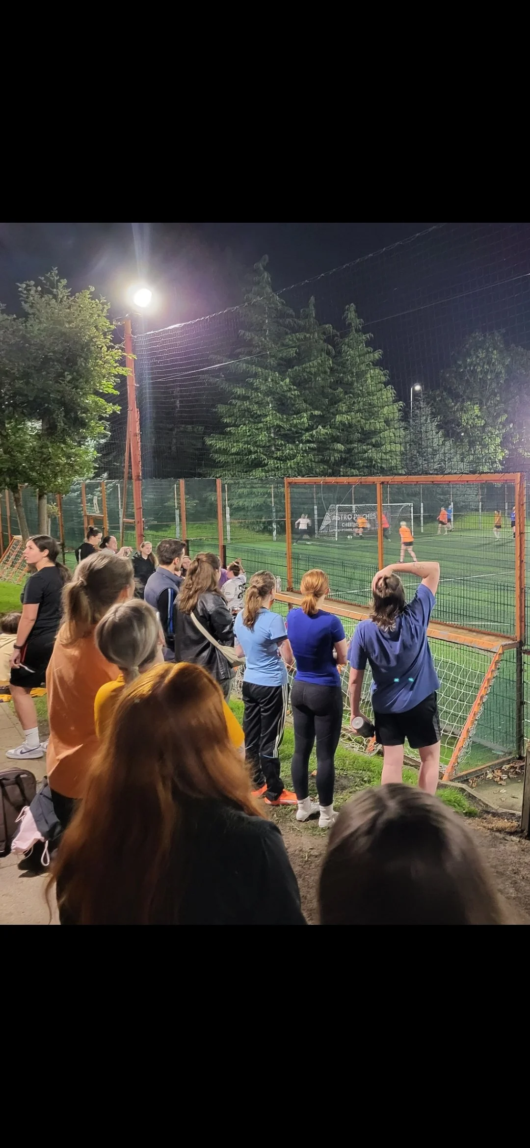 People watching a soccer game through a fence at night, on a grassy field surrounded by trees and a tall net.