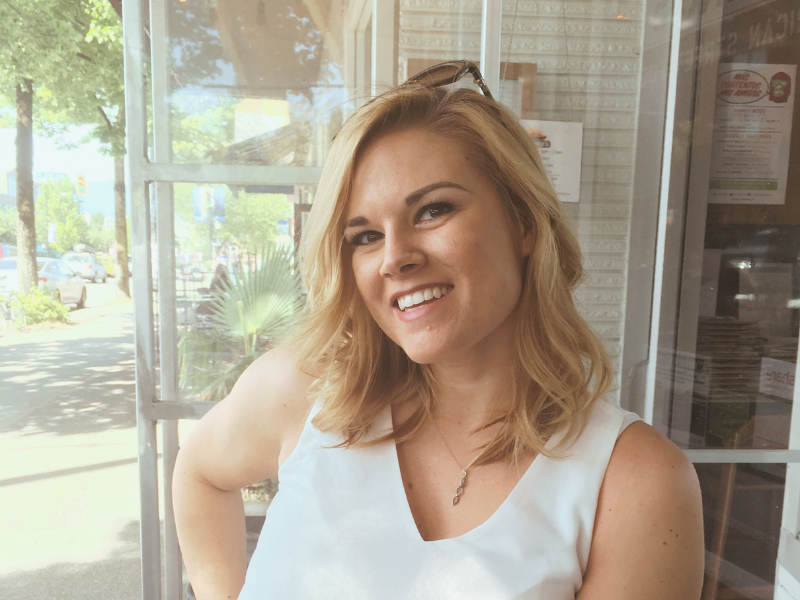 A young woman with shoulder-length blonde hair, wearing a white sleeveless top, smiling and standing inside a restaurant or cafe near a large window.