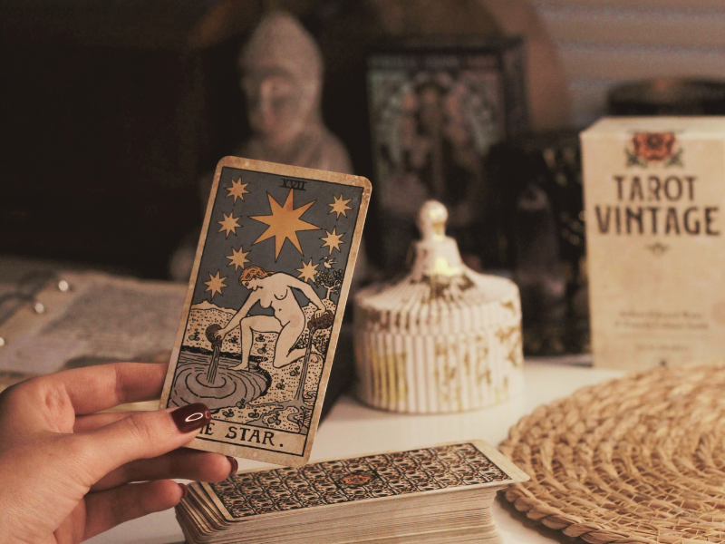 Person holding the tarot card 'The Star' in front of a desk with tarot cards, a vintage-style container, and tarot-themed items in the background.