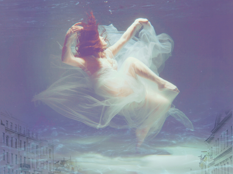 A woman wearing a white dress appears to be floating or swimming above a city street reflected on water.