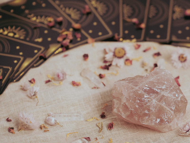 A large pink crystal, tarot cards, and flower petals on a white cloth surface.