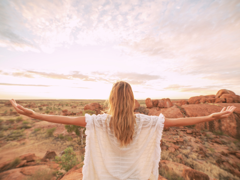 A woman with long red hair standing on rocky desert terrain with arms outstretched, facing a sunset sky with scattered clouds.