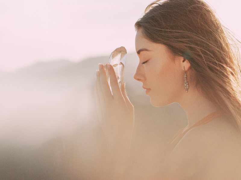 A woman with long brown hair and earrings holding a crystal or mineral to her forehead, eyes closed, outdoors at sunset with a soft, blurred background.