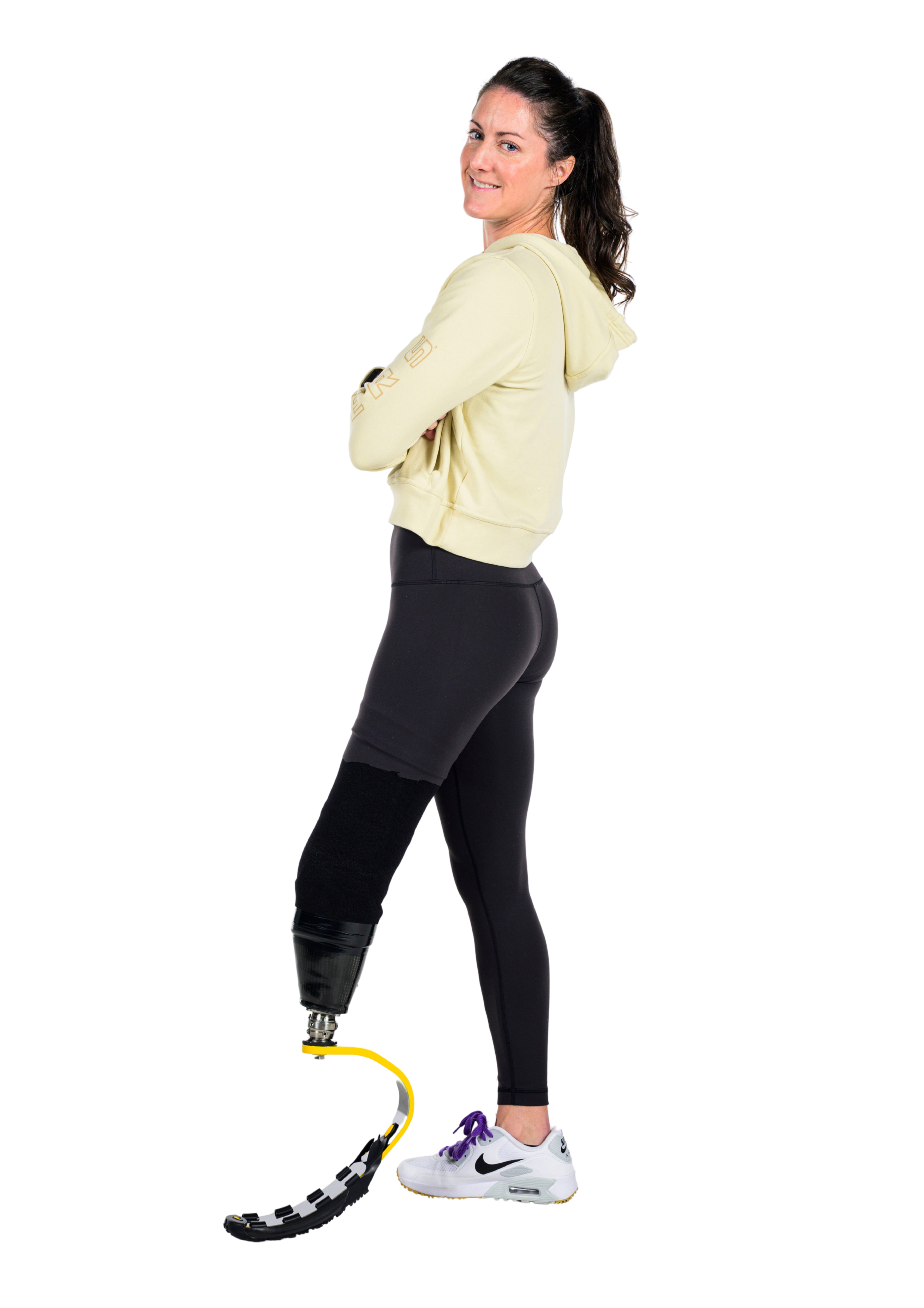 A woman with dark hair in a high ponytail, wearing a cream hoodie and black leggings, standing on a wooden floor. She has a prosthetic leg and is wearing white Nike sneakers, with a black and purple athletic prosthetic foot, and a yellow taping on her left foot. She is smiling and looking over her shoulder.