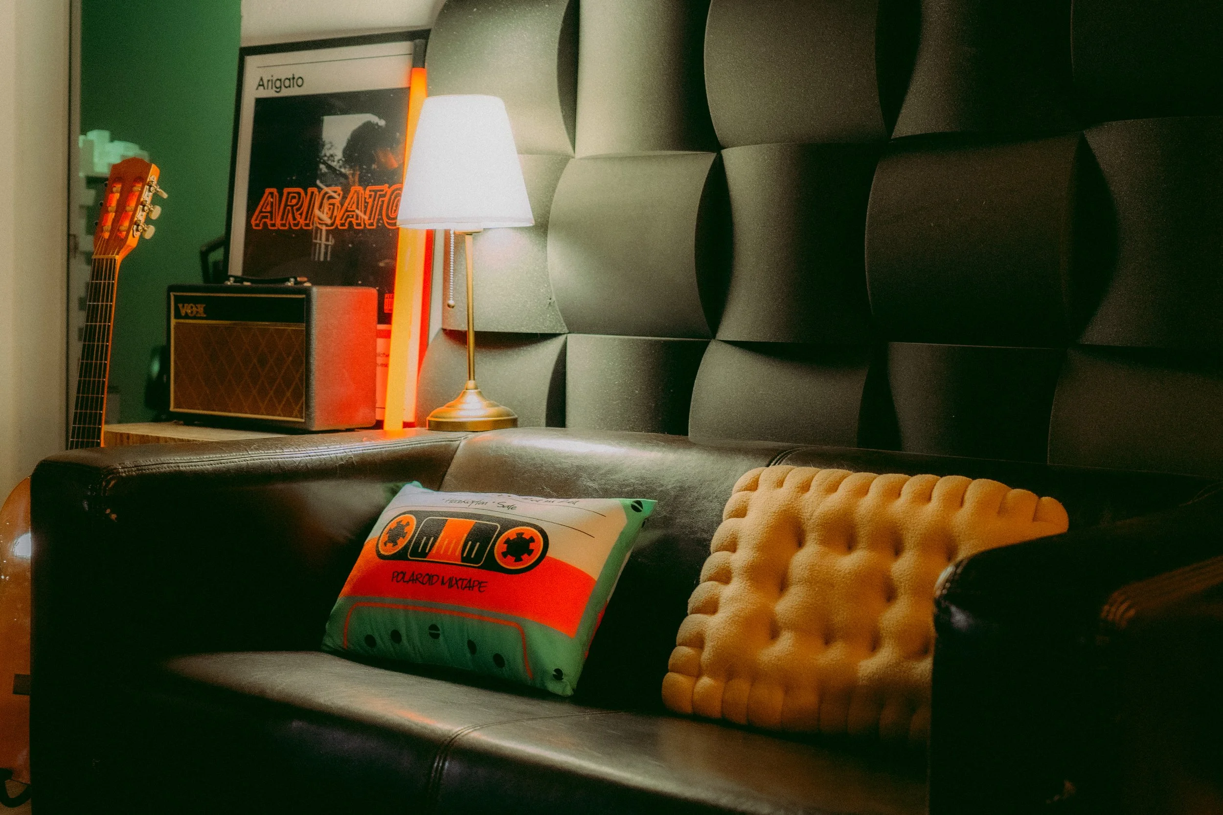 Eine Wohnzimmerecke mit einem schwarzen Ledersofa, einem gelben Kissen mit Struktur, einem Kissen, einem kleinen Gitarrenverstärker, einer weißen Lampe, einem gerahmten Bild mit der Aufschrift „Arigato“ und einer Wand mit grauer Textur.