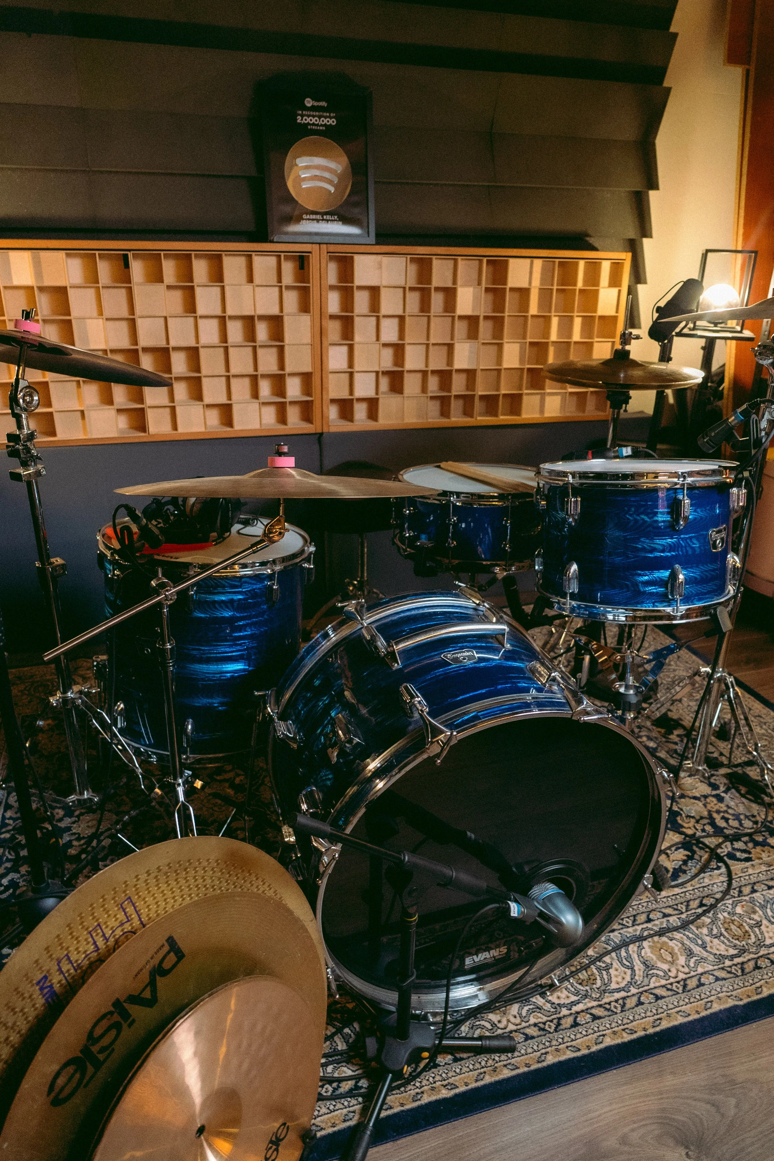 Ein Musikaufnahmestudio mit einem blauen Schlagzeug auf einem gemusterten Teppich, Akustikplatten an der Wand, einem Musikposter und einem im Vordergrund teilweise sichtbaren Becken.
