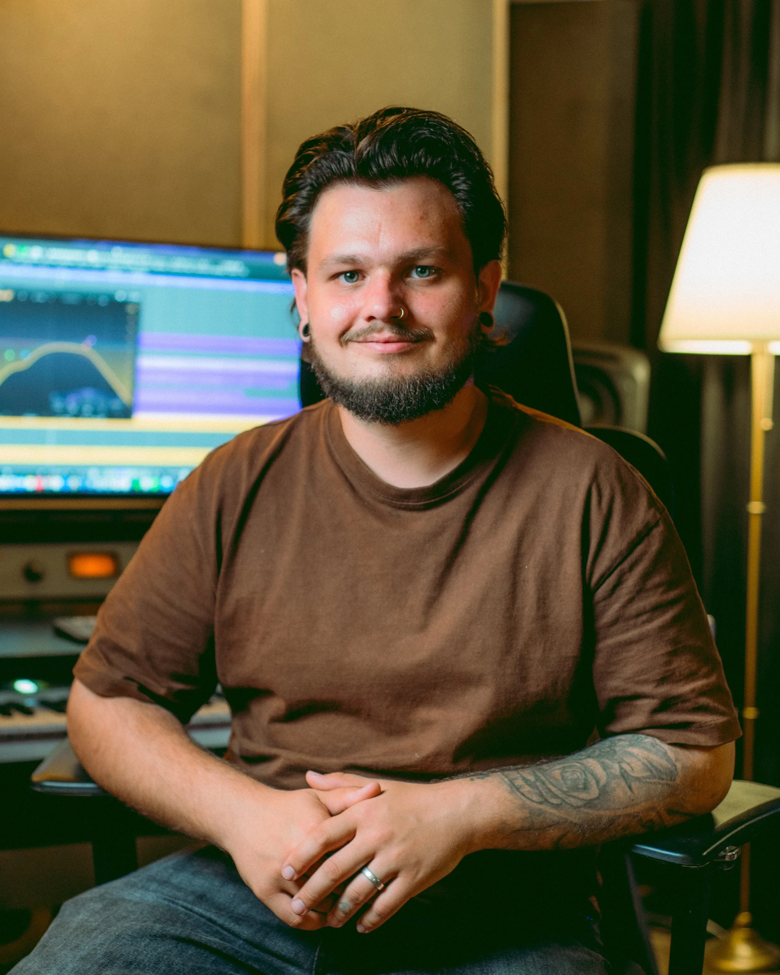 Eine Person mit heller Haut, blauen Augen und nach hinten gekämmten dunklen Haaren, die in einem Aufnahmestudio oder einem Musikproduktionsraum sitzt, ein braunes T-Shirt trägt, Tätowierungen am linken Arm, Ringe an den Fingern und Ohrringe hat