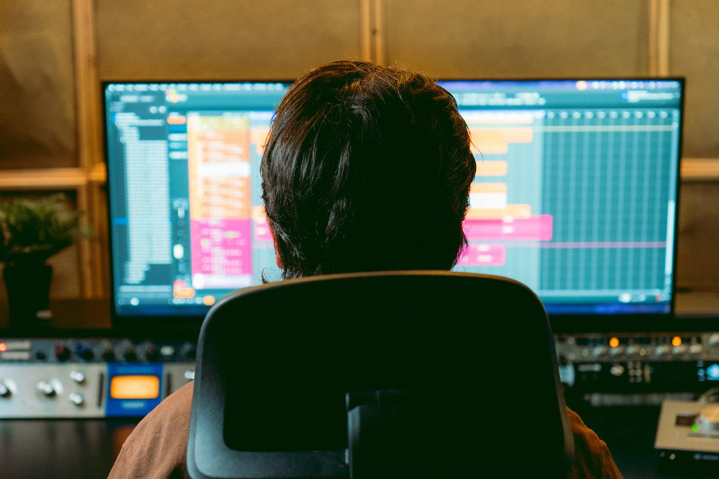 Eine Person mit dunklen Haaren, die in einem Tonstudio an einem Computer arbeitet, auf dessen Bildschirm die Benutzeroberfläche einer großen Musikproduktionssoftware zu sehen ist.