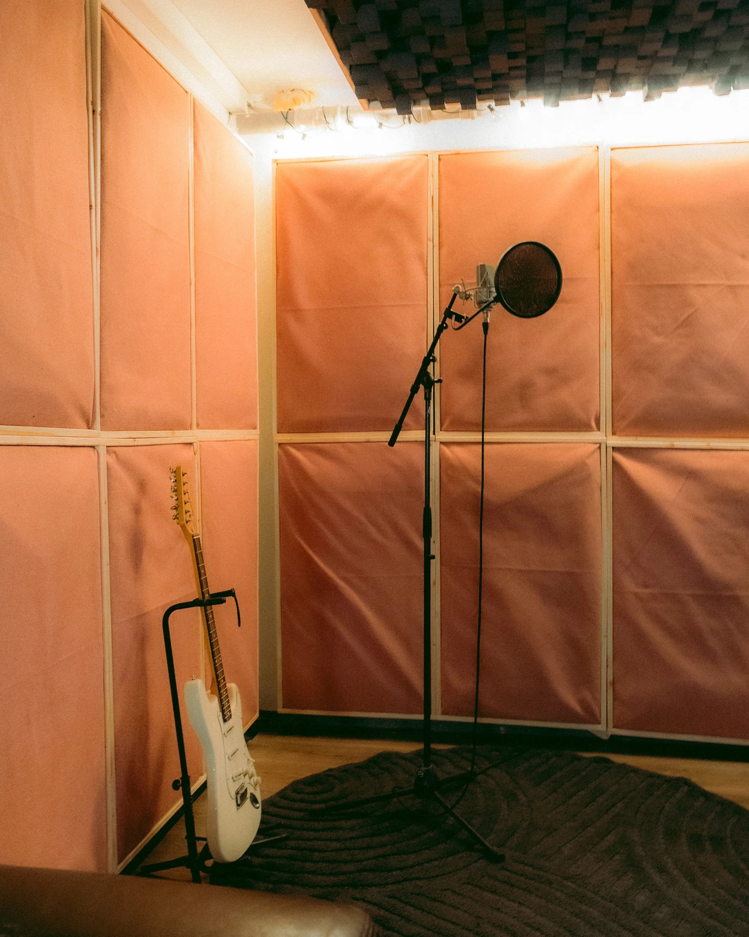 Aufnahmestudio mit rosa Akustikplatten, einem Mikrofon auf einem Ständer und einer E-Gitarre auf einem Ständer, dazu ein schwarzer Teppich auf einem Hartholzboden.