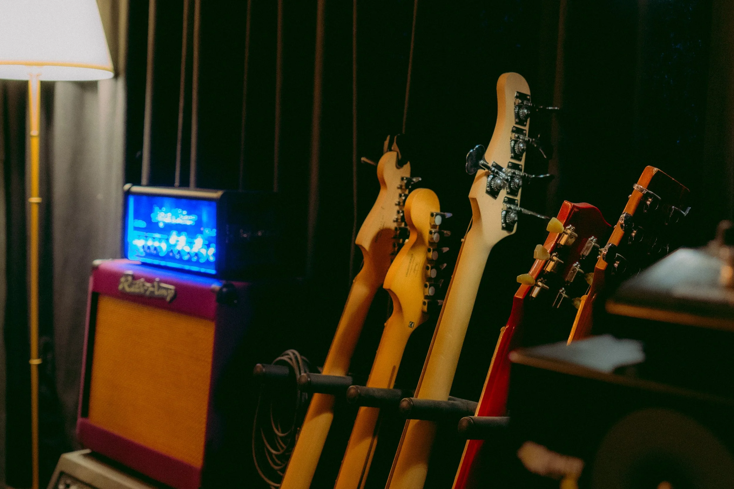 Eine Reihe von E-Gitarren in verschiedenen Farben und Ausführungen, die aufrecht an einer dunklen Wand stehen, flankiert von bunten Gitarrenverstärkern und einem neonblauen Schild im Hintergrund.