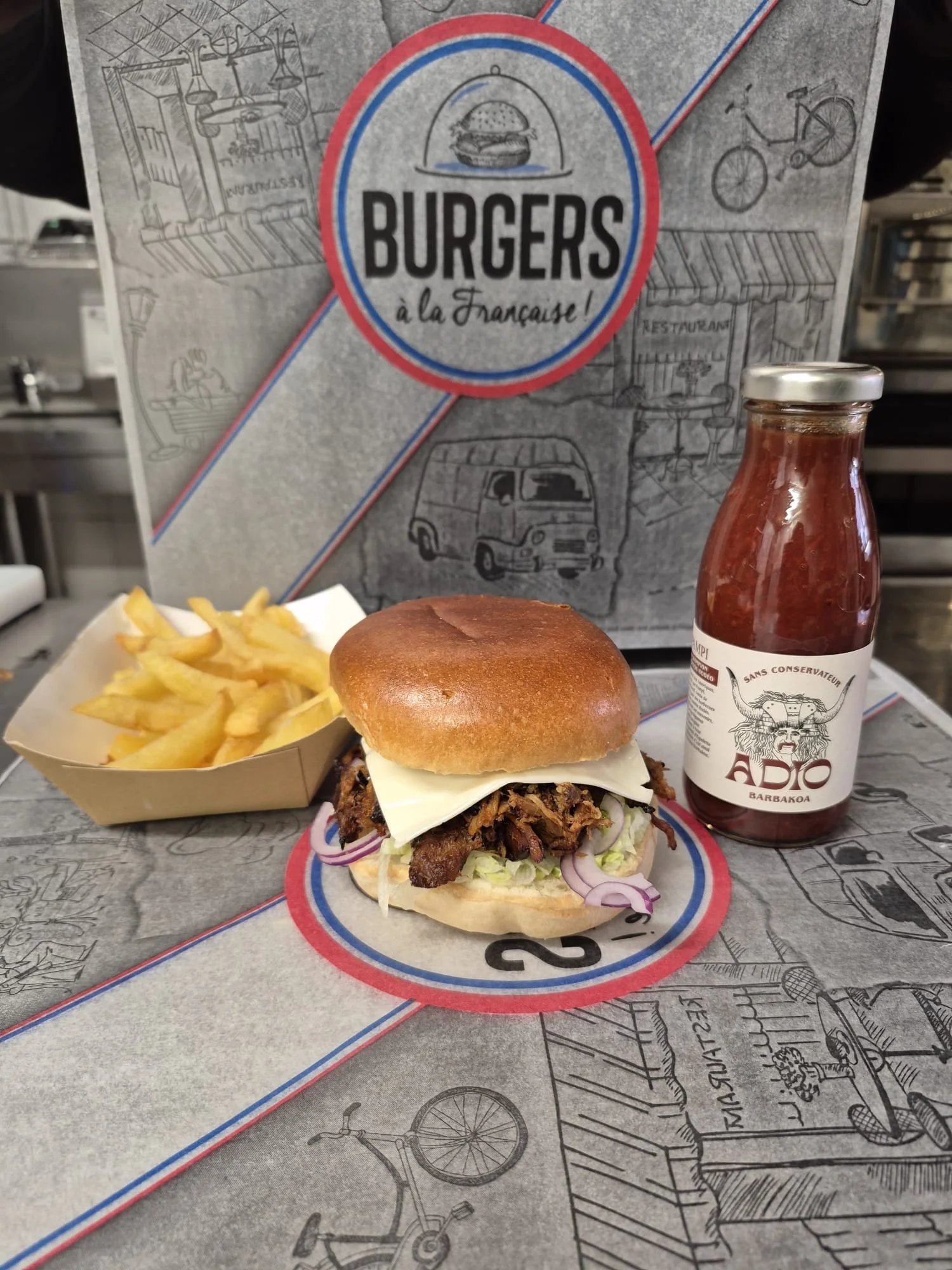 Un burger avec de la viande, du fromage, de la laitue et des oignons, accompagné de frites, une bouteille de ketchup et une boisson dans une bouteille en verre. Le fond montre un panneau avec le texte "Burgers à la Française".