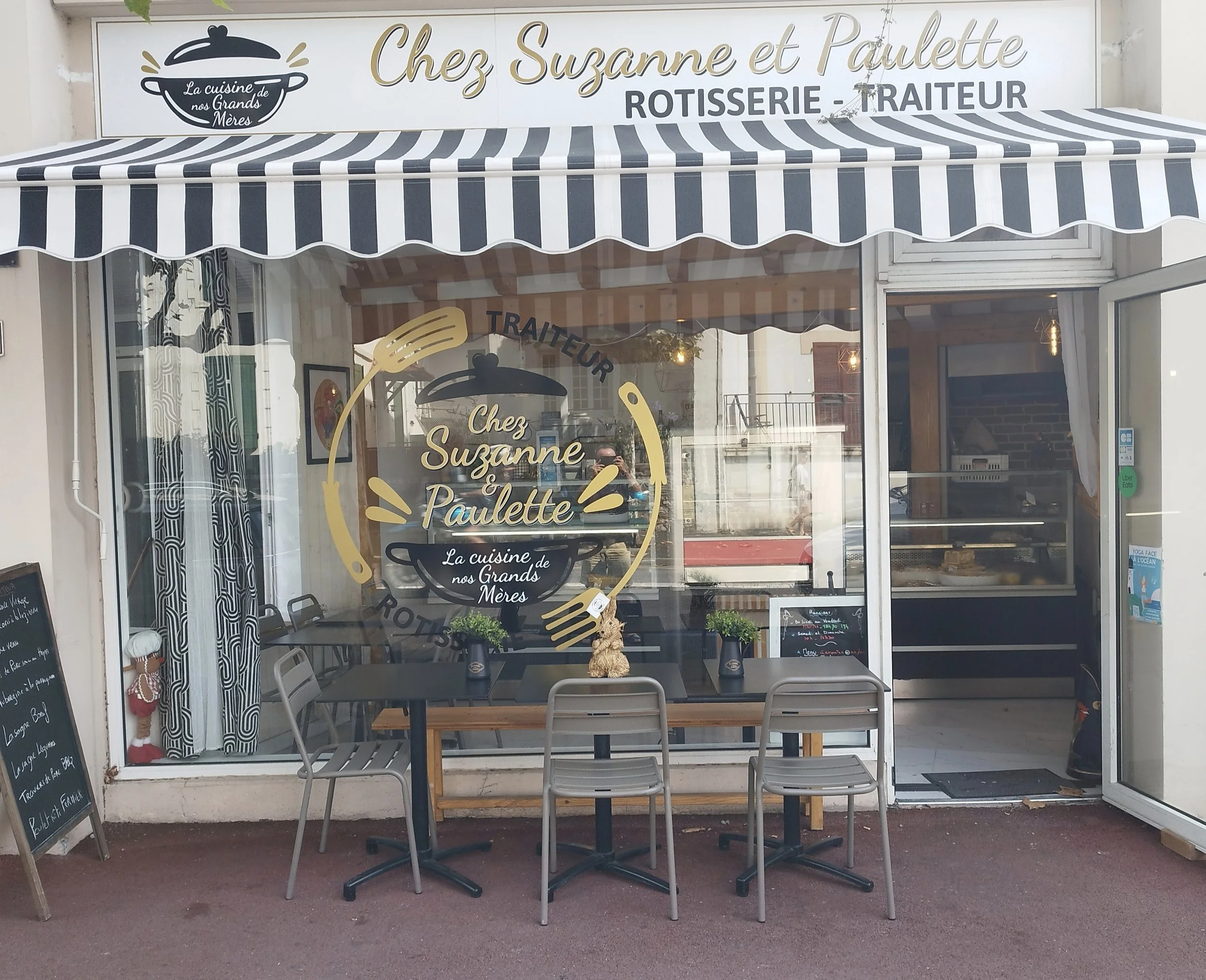 Façade d'un restaurant appelé 'Chez Suzanne et Paulette' avec une enseigne indiquant 'Rotisserie - Traiteur'. Il y a une vitrine, une table avec des chaises, et un panneau noir à gauche avec le menu. Il y a aussi des décorations comme une petite poupée et des plantes à l'intérieur.