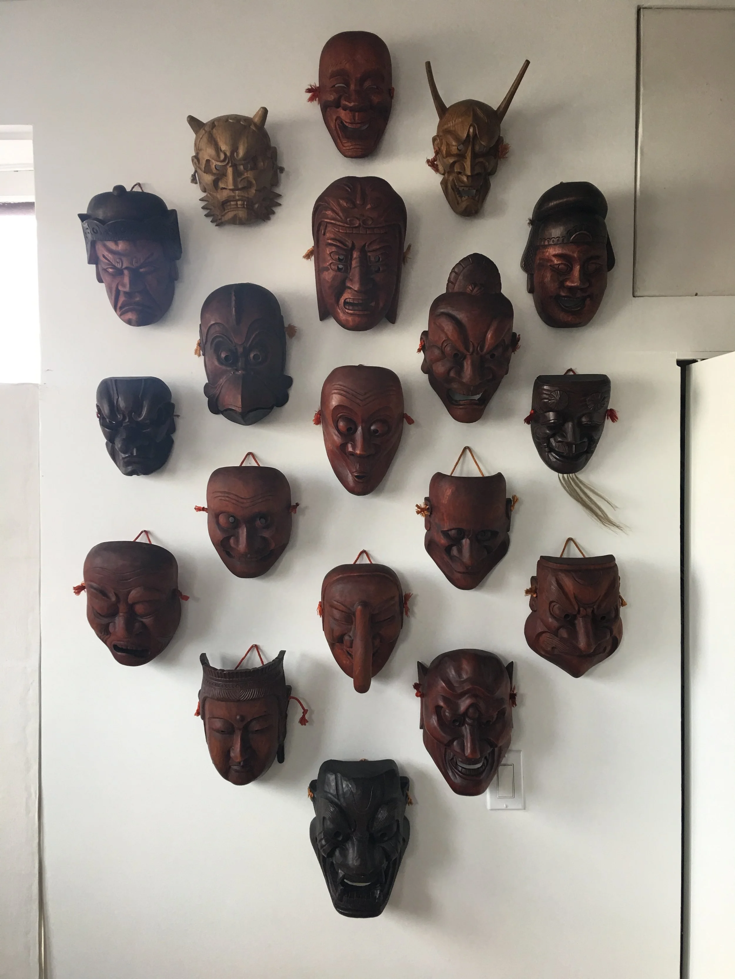 Wall display of various traditional wooden masks, arranged in rows on a plain white wall.
