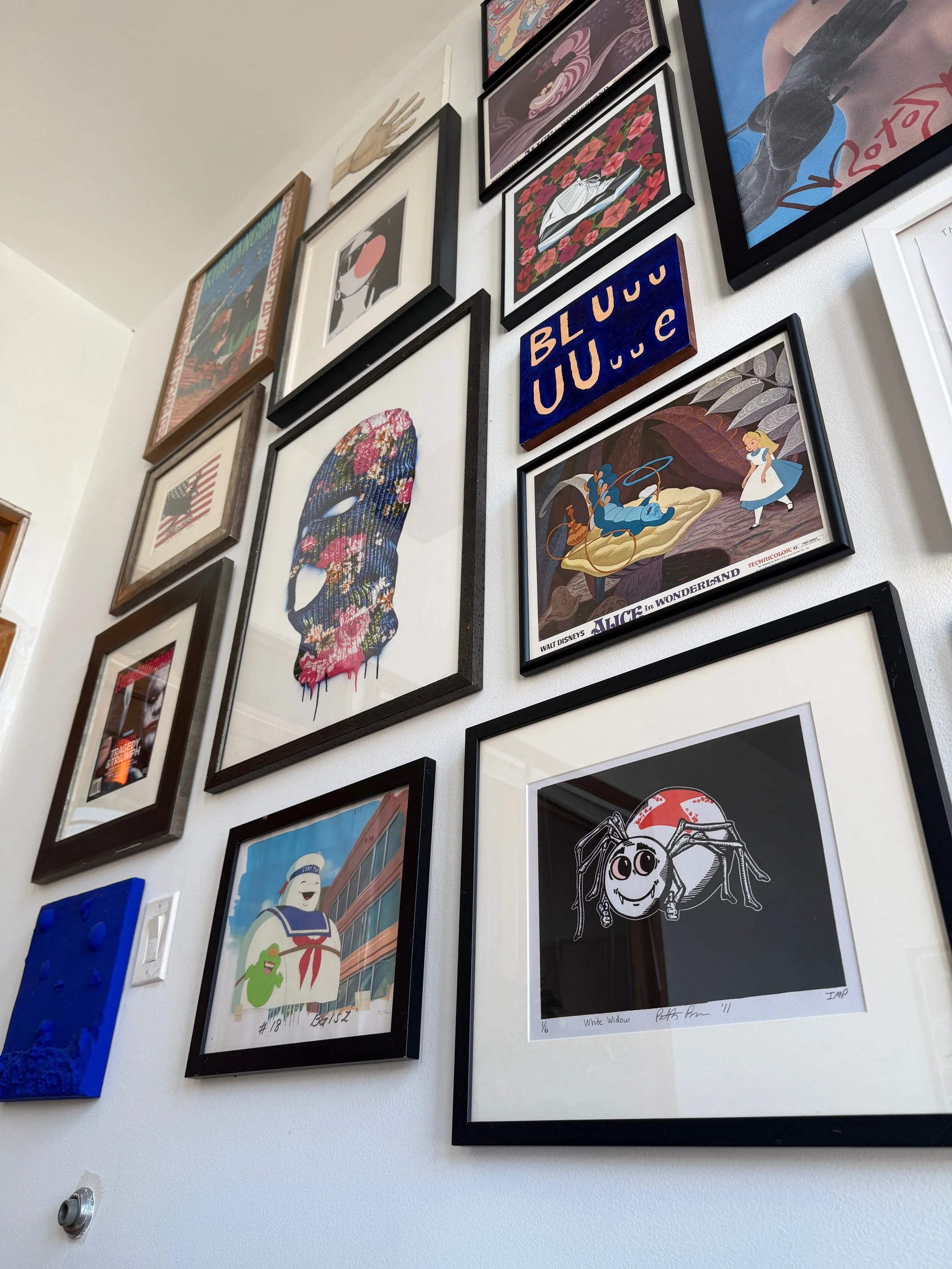 Gallery wall with various framed artwork including illustrations, posters, and paintings. Artwork includes a colorful skull, Alice in Wonderland scene, a Spider-Man poster, and various abstract and pop-culture pieces.
