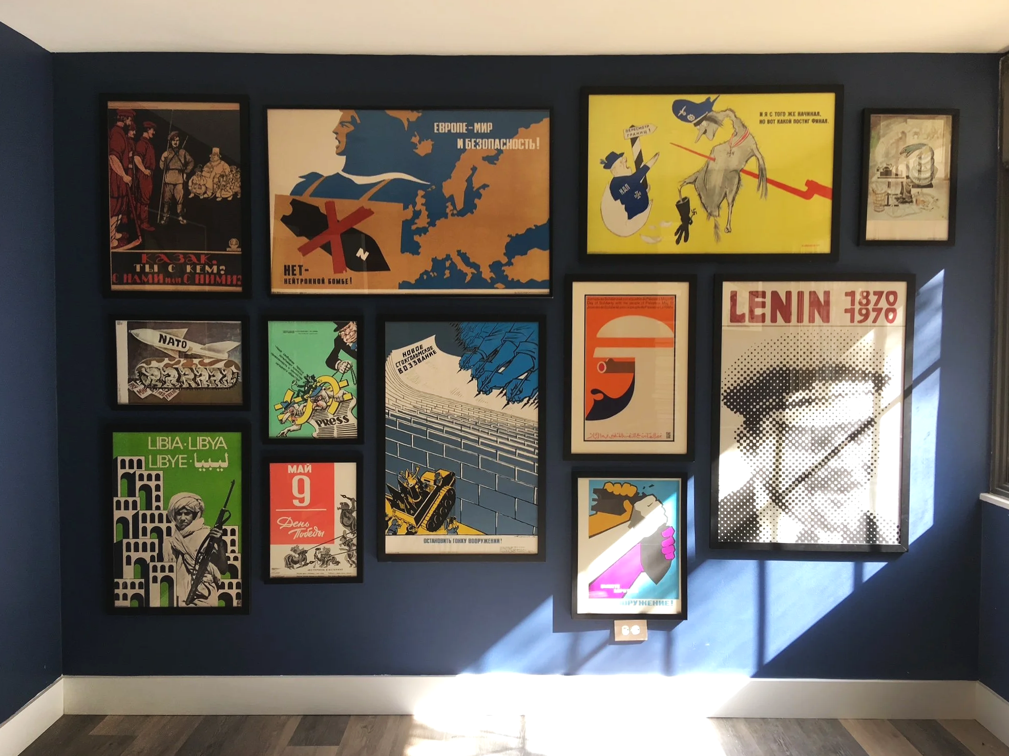 A wall display of framed propaganda posters, including images related to NATO, Russia, and Lenin, arranged on a dark blue wall with lighting overhead.