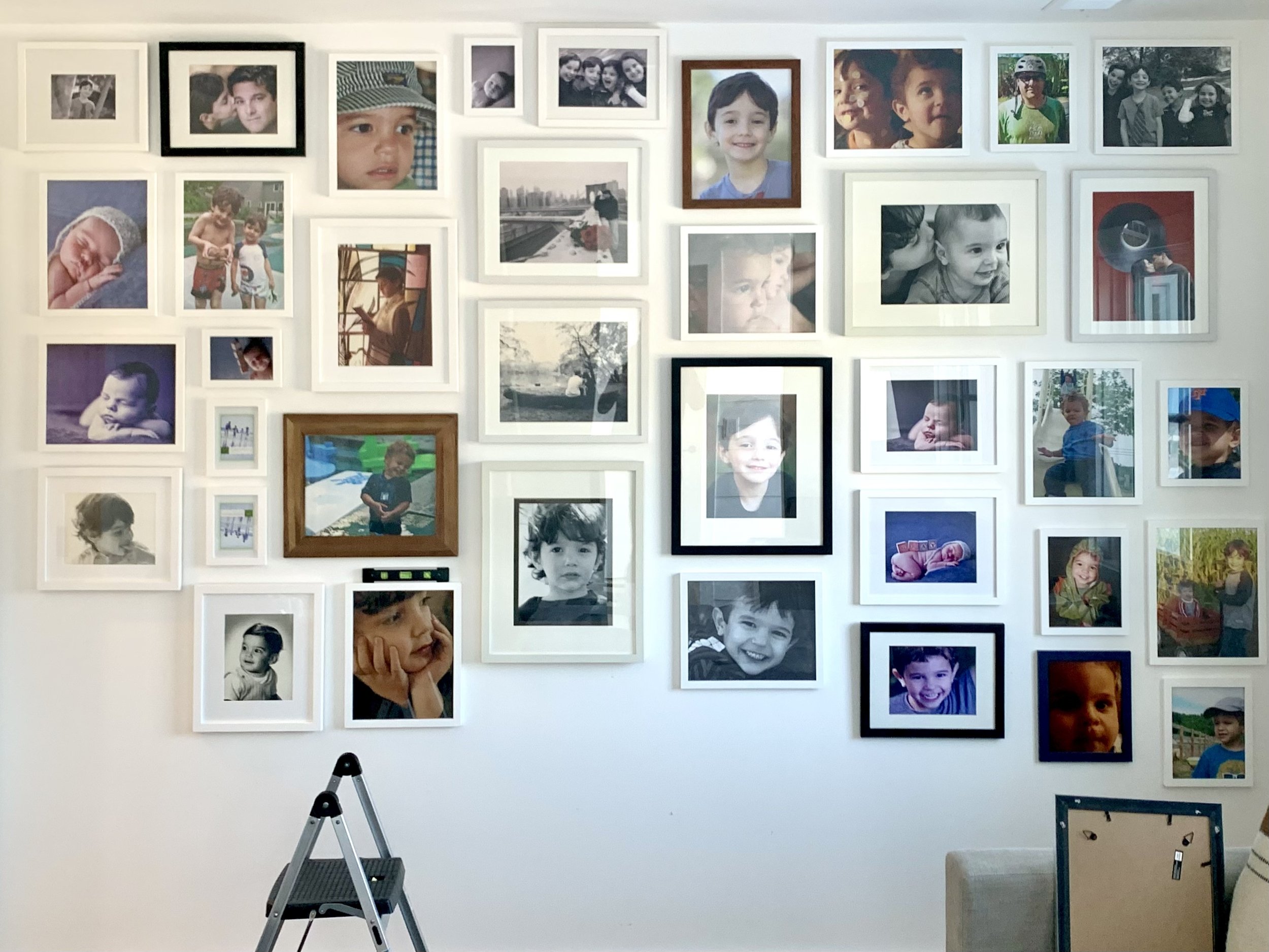 A wall filled with numerous framed photographs of children and family moments. The photos include children playing, smiling, and various outdoor activities. Some photos are black and white, while others are in color. There are two step ladders, one i