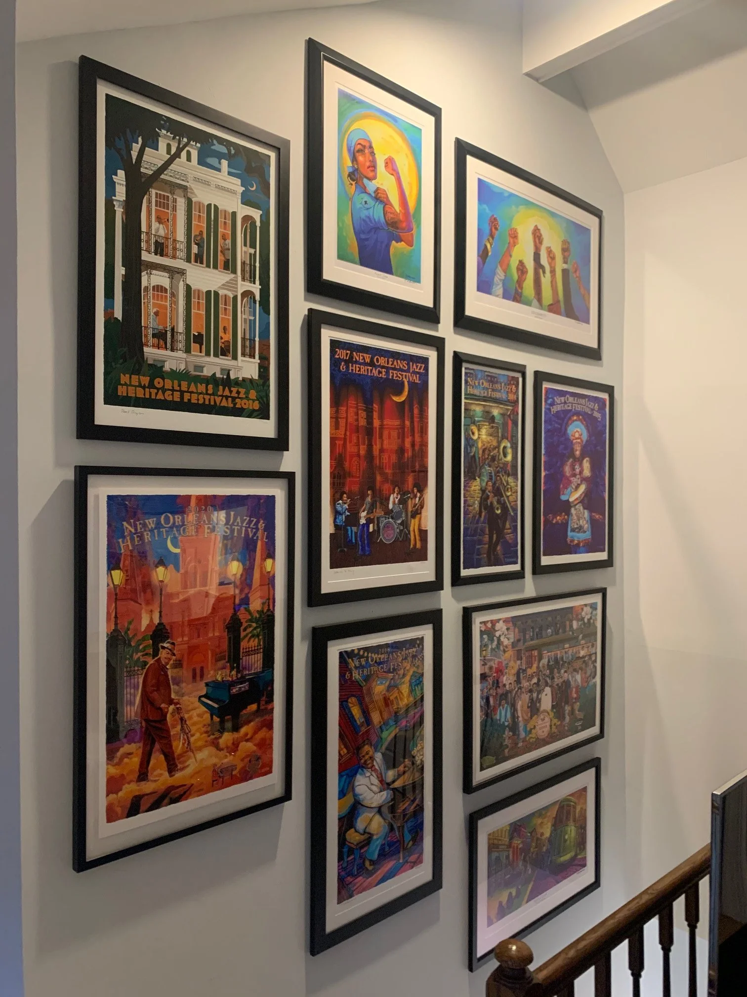 A gallery wall on a stairwell featuring colorful posters of the New Orleans Jazz & Heritage Festival from various years, arranged in black frames.