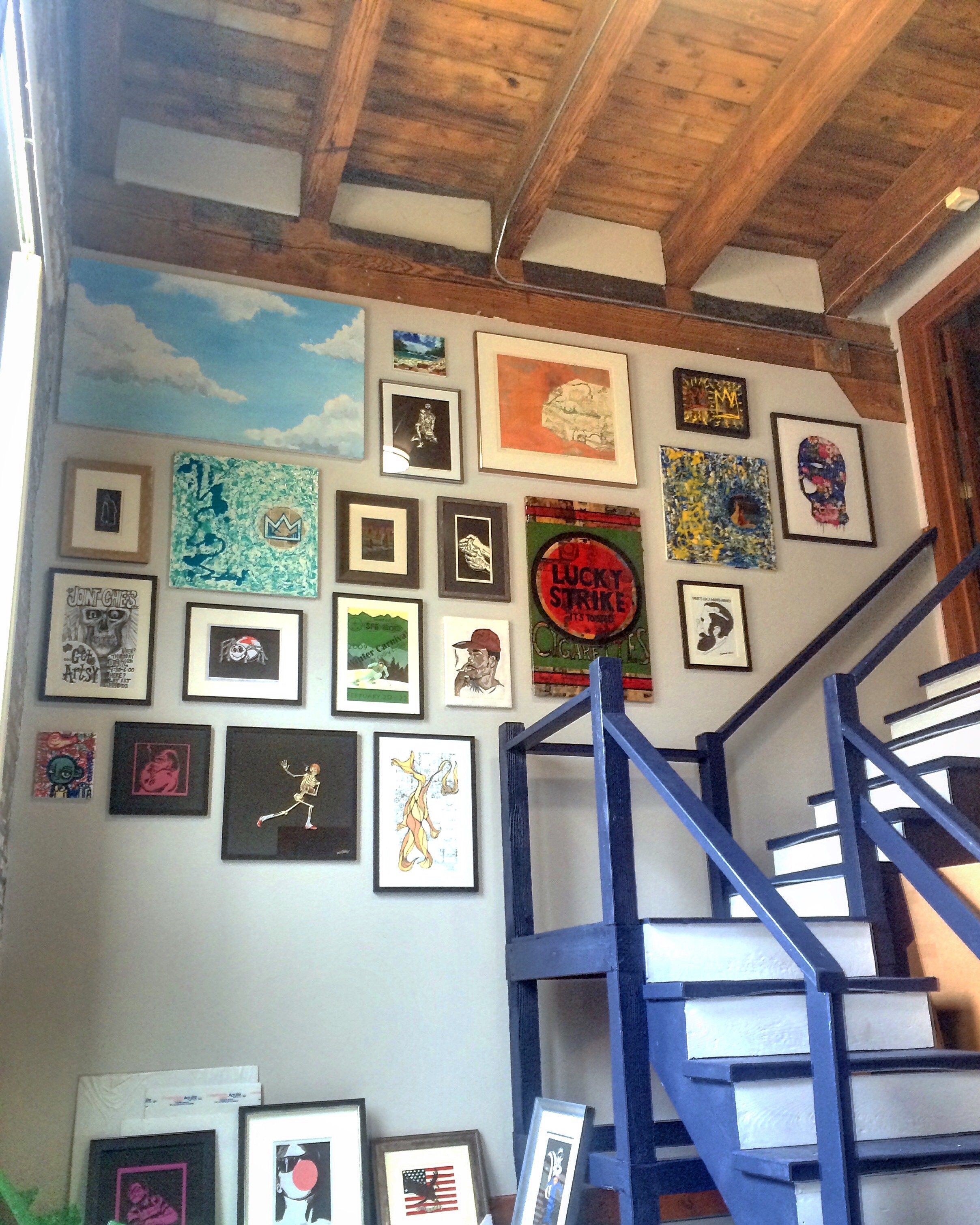 Interior view of an art gallery or studio with wall decorated with various framed artwork. The artwork includes paintings, prints, and illustrations, some with recognizable themes like clouds and abstract designs. There's a wooden ceiling with expose