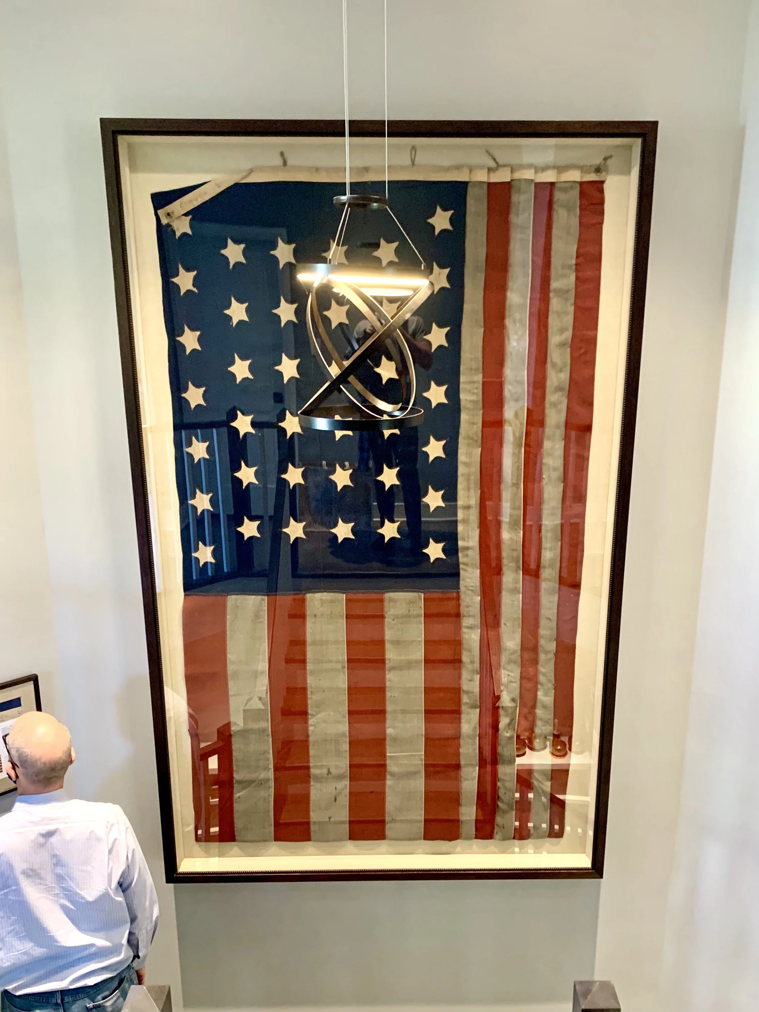 Large American flag artwork displayed vertically on a wall behind a person with a bald head and white shirt, in a museum or gallery setting.