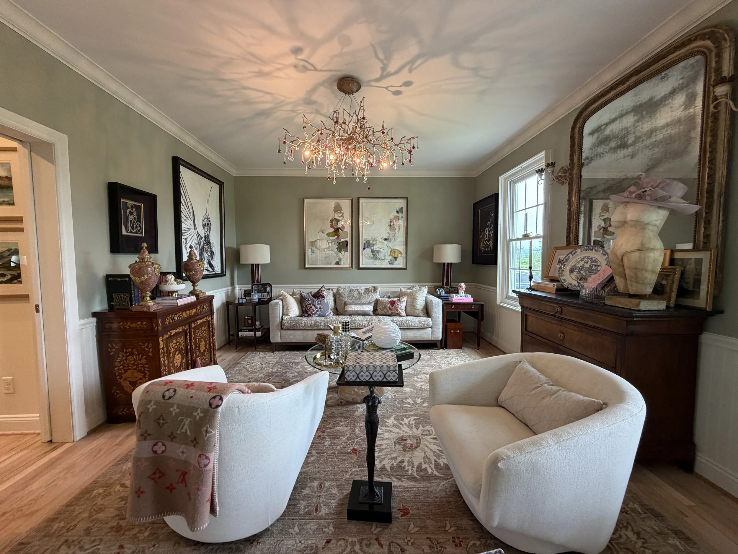 Living room with beige sofa, white armchair with a patterned cover, a glass coffee table, artwork on the walls, and a chandelier casting decorative shadows on the ceiling.