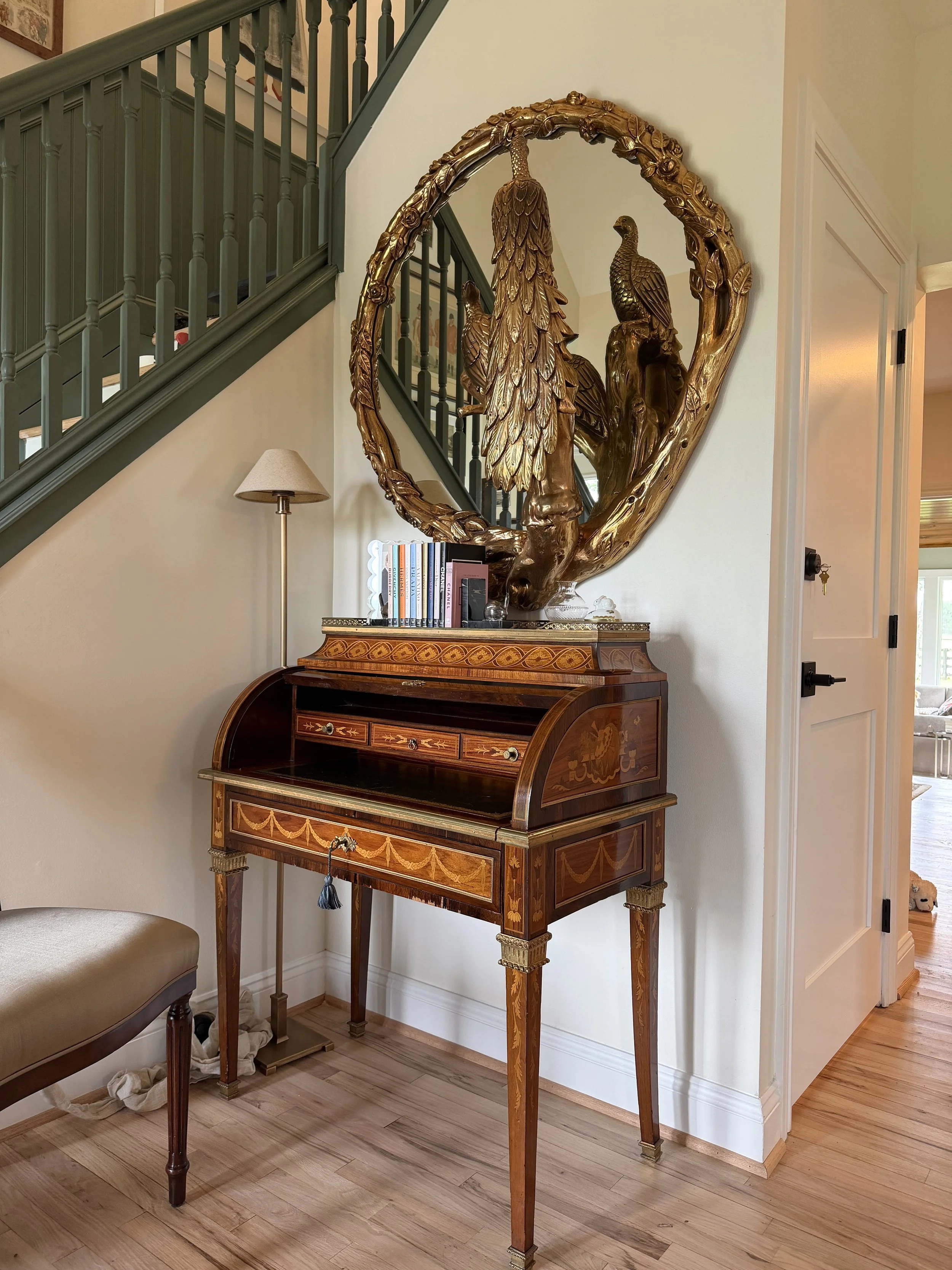 A wooden secretary desk with inlaid marquetry design, topped with a decorative gold-colored sculpture of birds surrounded by flowers and leaves, located against a wall with a staircase in the background.