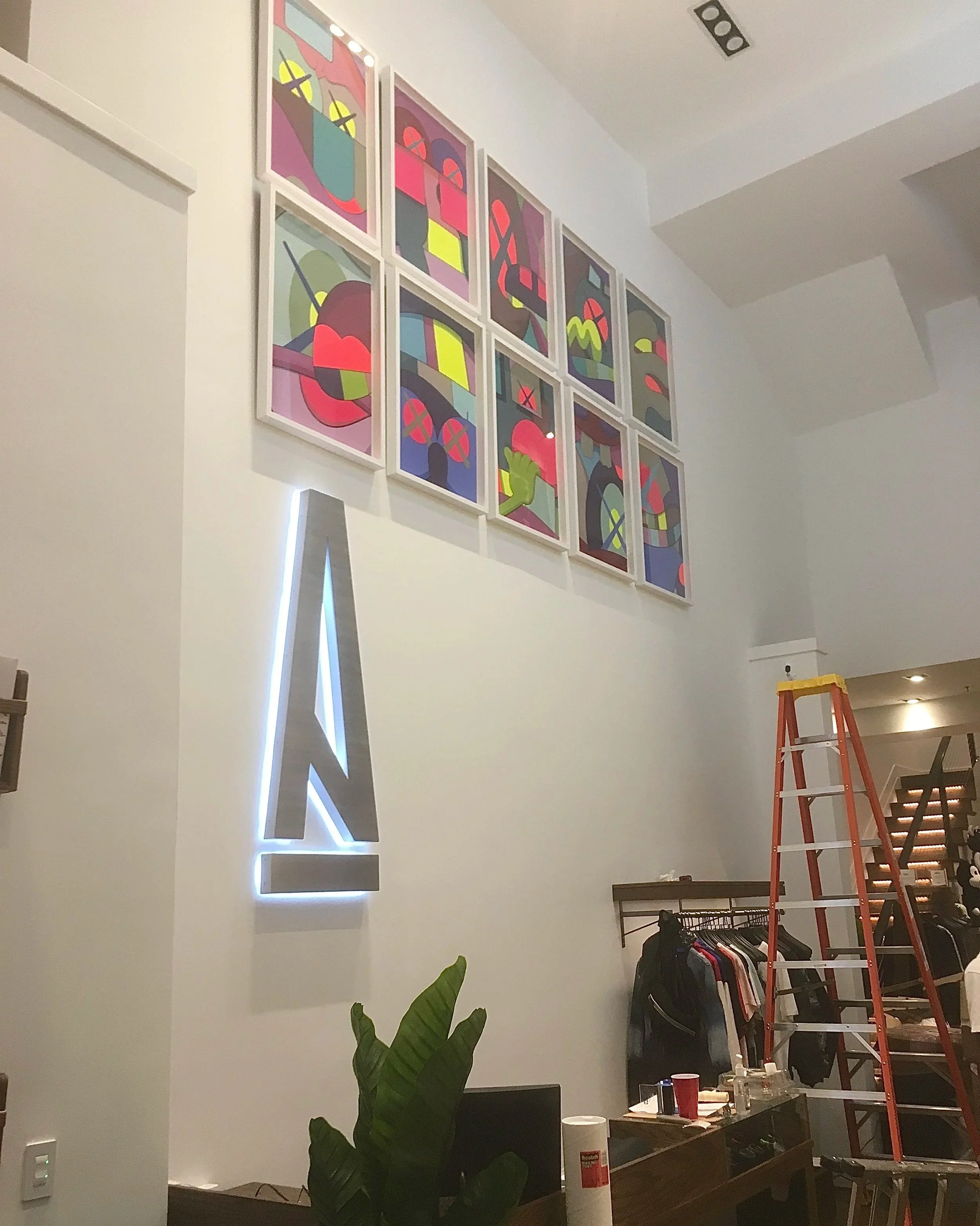 Photo of a tall wall decorated with a set of colorful abstract paintings arranged in a grid. A ladder and various household items are in the room, which appears to be under setup or organization.