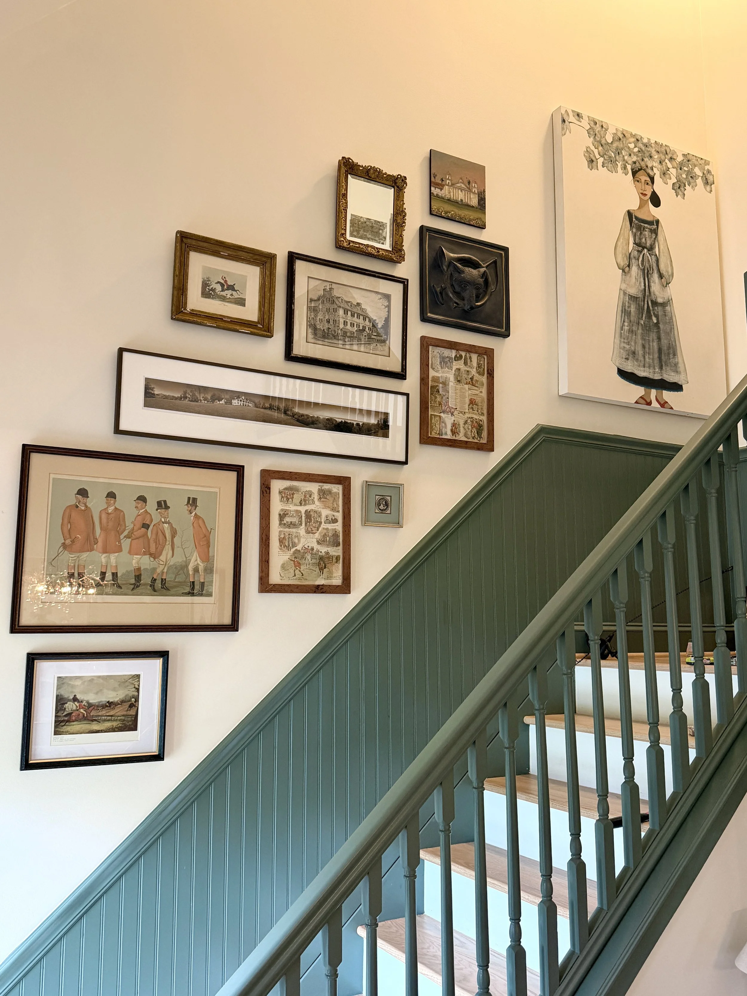Wall decorated with various framed artworks and paintings, including portraits, landscapes, and historical illustrations, with a staircase painted green on the right side.