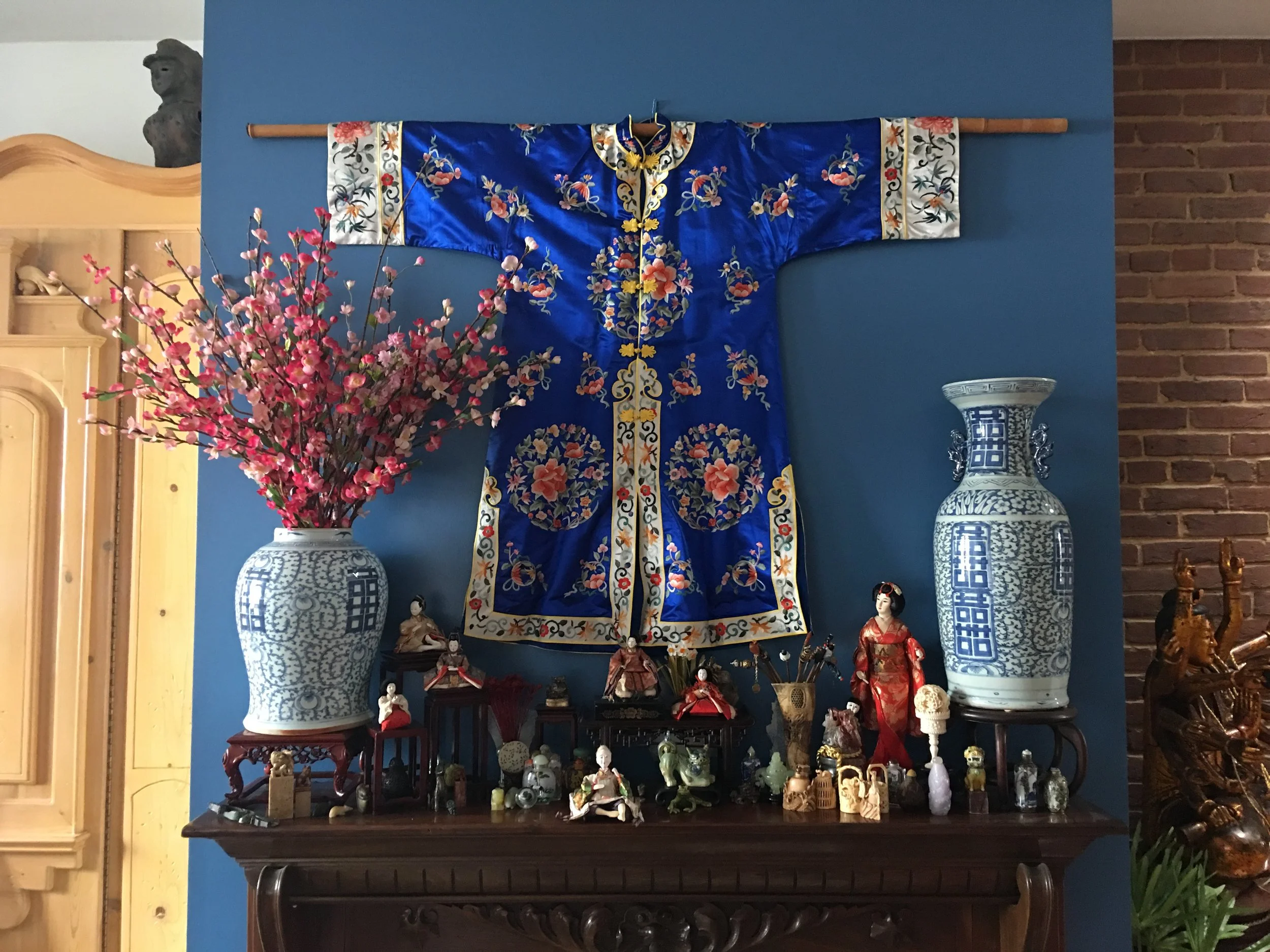 An Asian-themed display on a wooden table featuring various figurines, small sculptures, and decorative items. Behind it, against a blue wall, are large blue and white porcelain vases, a pink flower arrangement, a decorative fabric with floral designs hanging on the wall, and additional cultural artifacts including puppet dolls and other decorative objects.