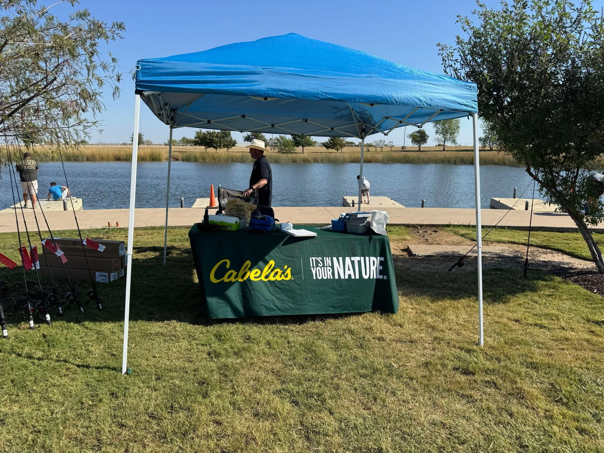 A fishing event setup at a lakeside with a blue canopy tent, a table with Cabela's branding, fishing rods on the ground, and people fishing in the water.