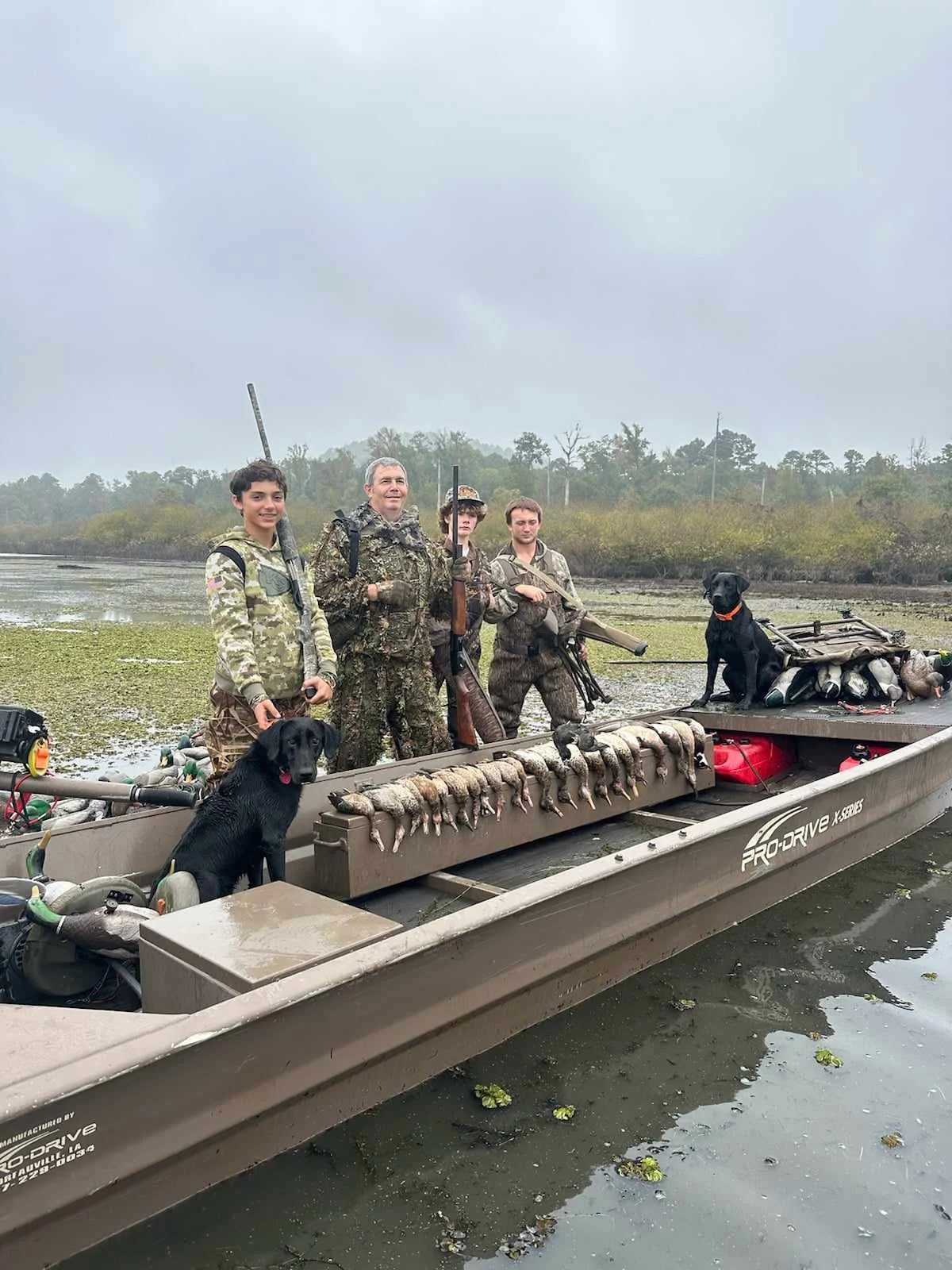 Four hunters with shotguns and two hunting dogs on a boat with ducks and decoys, on a marshy waterway on a cloudy day.