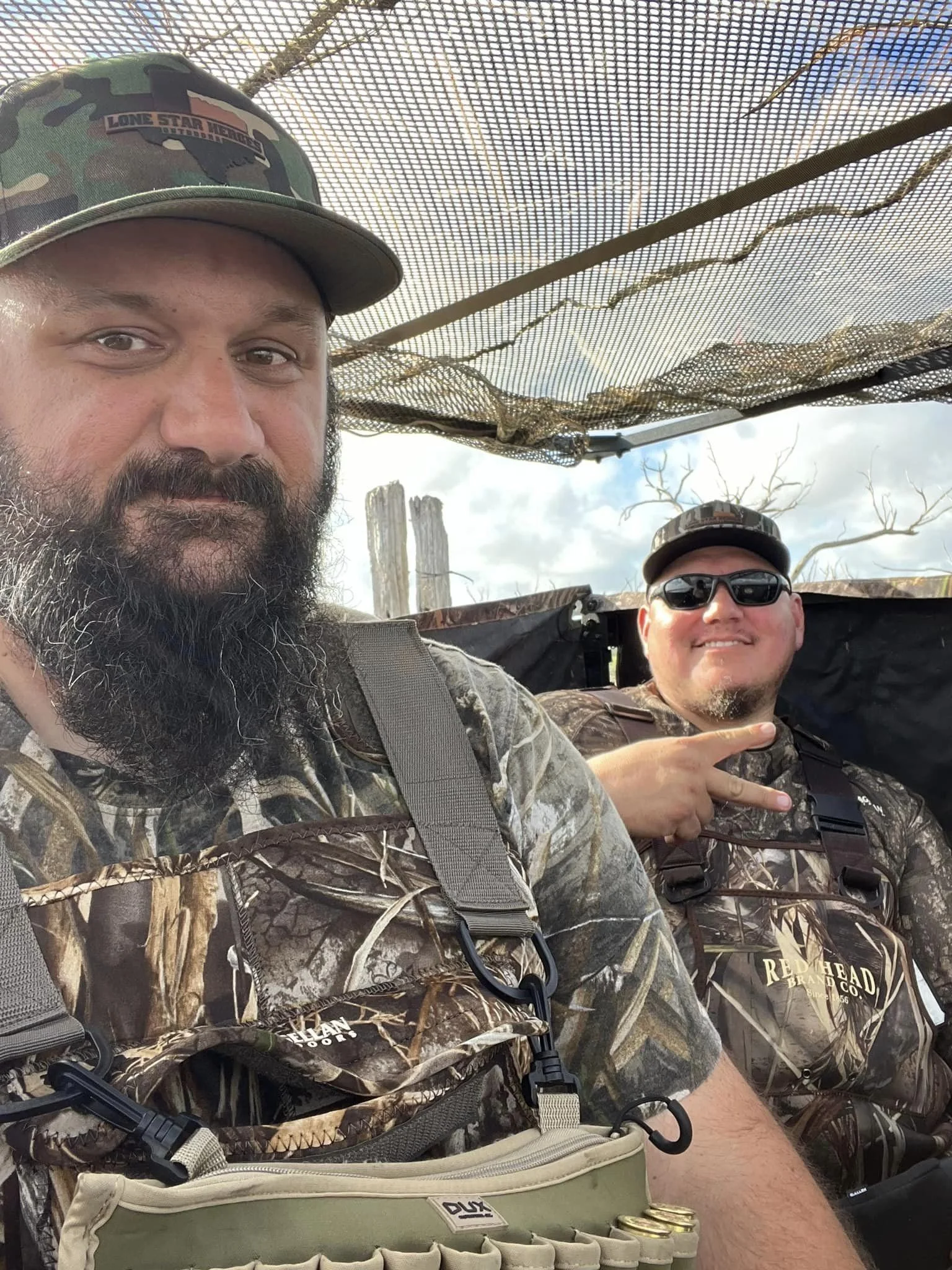 Two men in camouflage clothing sitting in a hunting blind, one making a peace sign, with a blue sky and trees in the background.