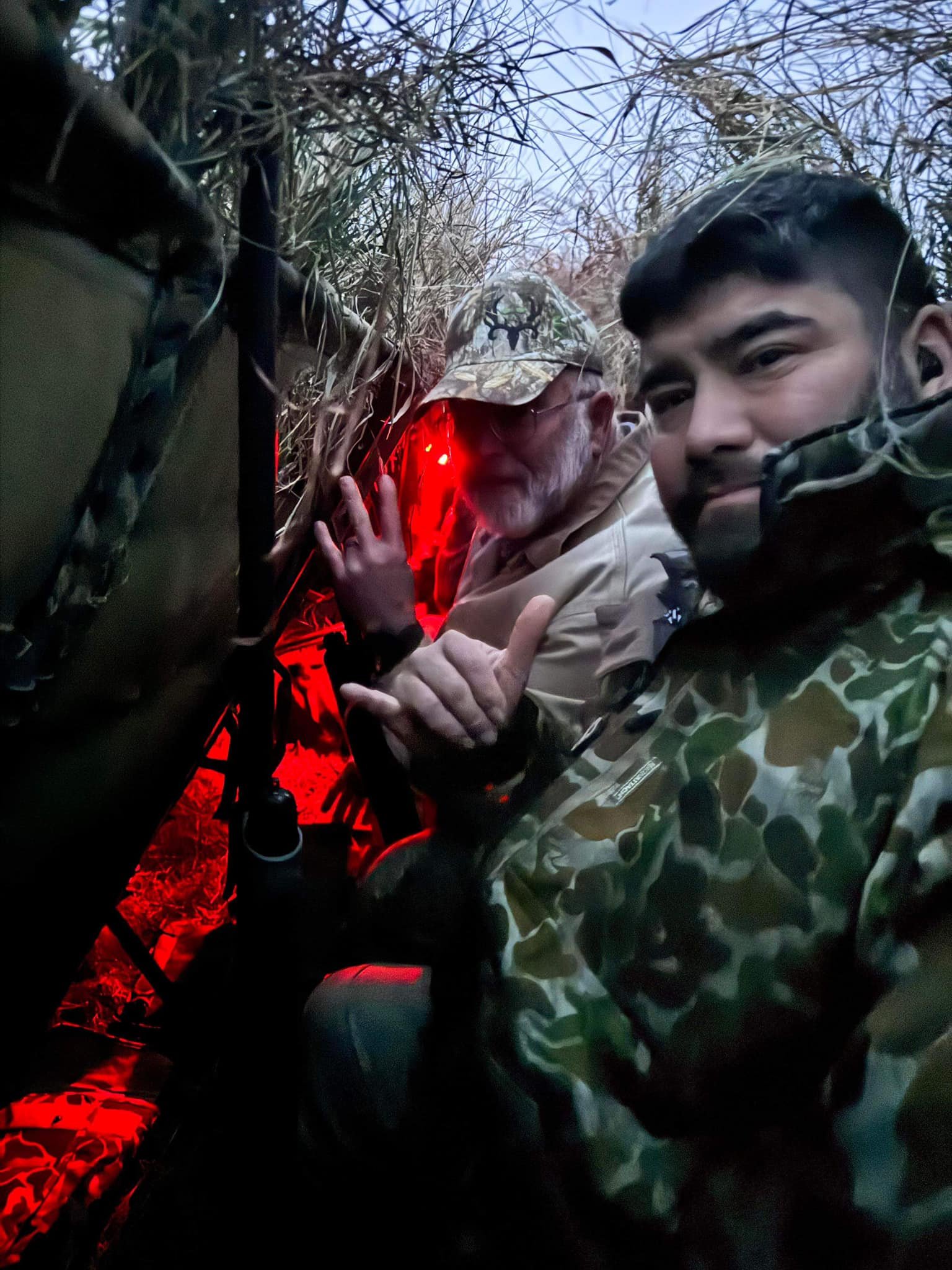 Two men in camouflage and tan clothing sitting in a narrow space surrounded by tall grass and bushes, with one man giving a thumbs-up and the other man resting against a dark object, both with serious expressions.