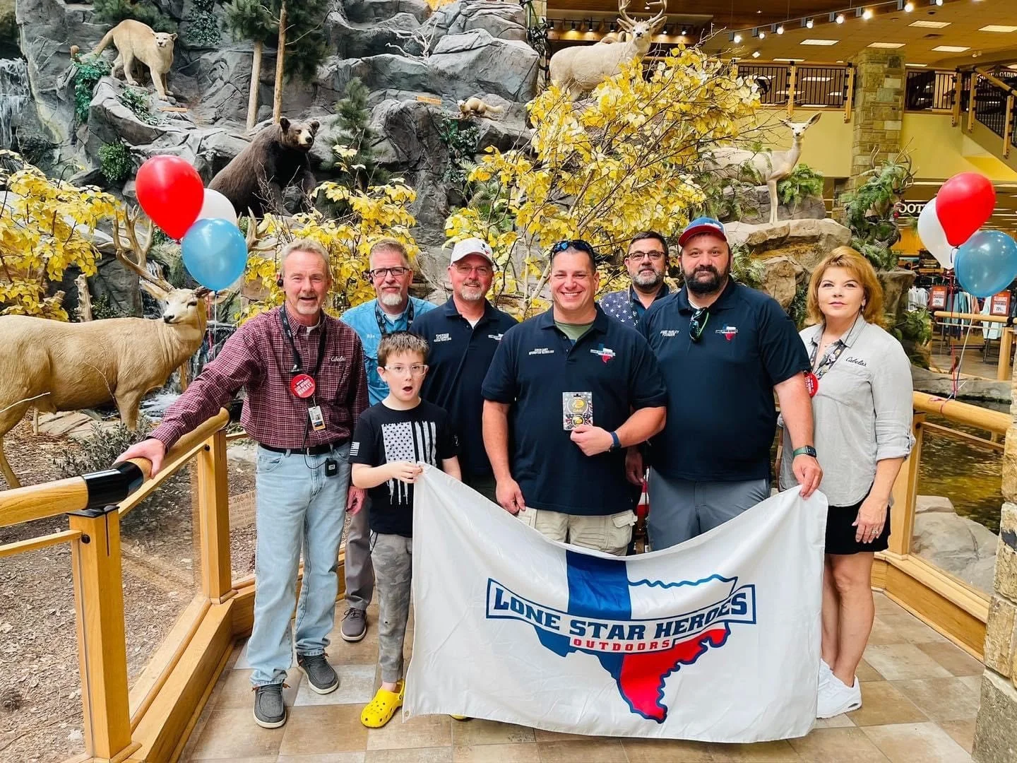 Group of eight people, including two children, holding a large "Lone Star Heroes Outdoors" flag, posing in an indoor wildlife exhibit featuring animals like deer, bear, and mountain lion, with artificial rocks and trees, and balloons.