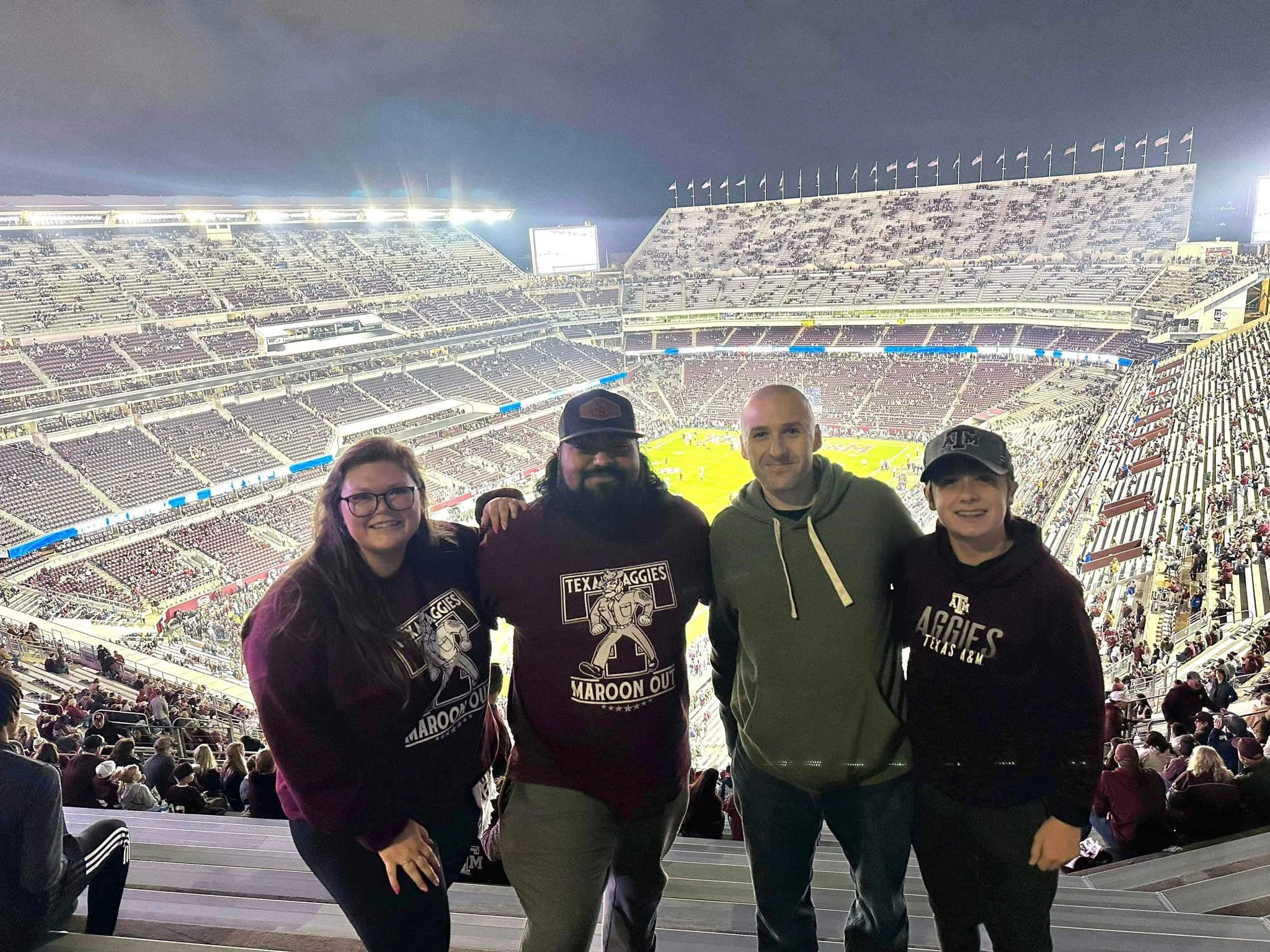 Four people standing together in a stadium during a night sports event, with the field and empty stands in the background.