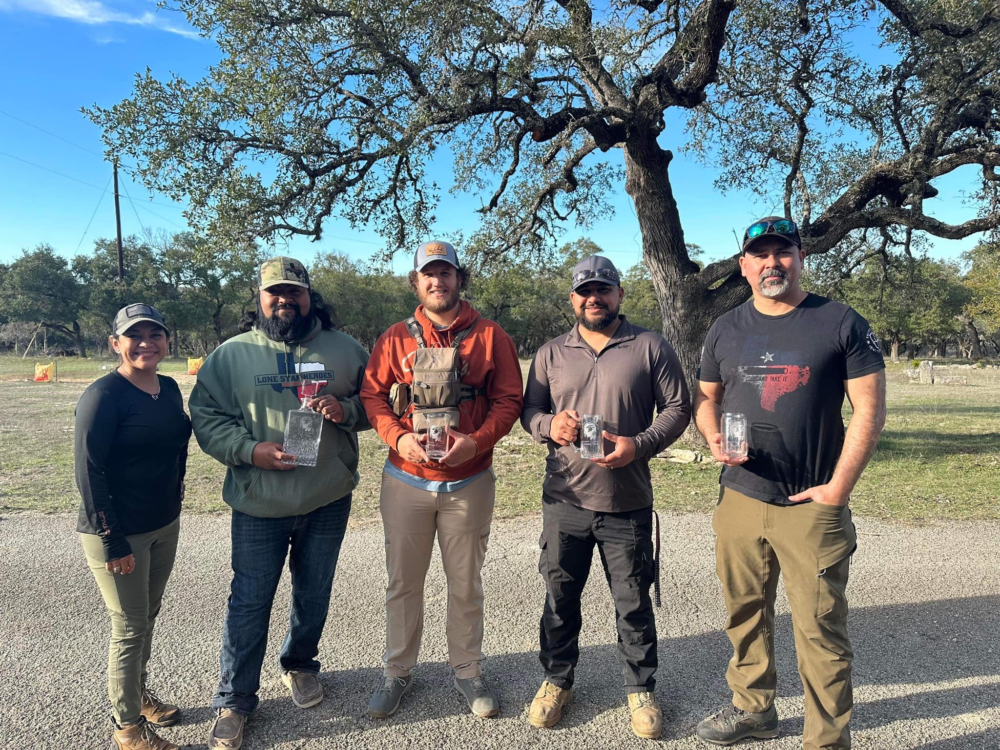 Five people standing outdoors on a sunny day, holding glass awards, with trees in the background.