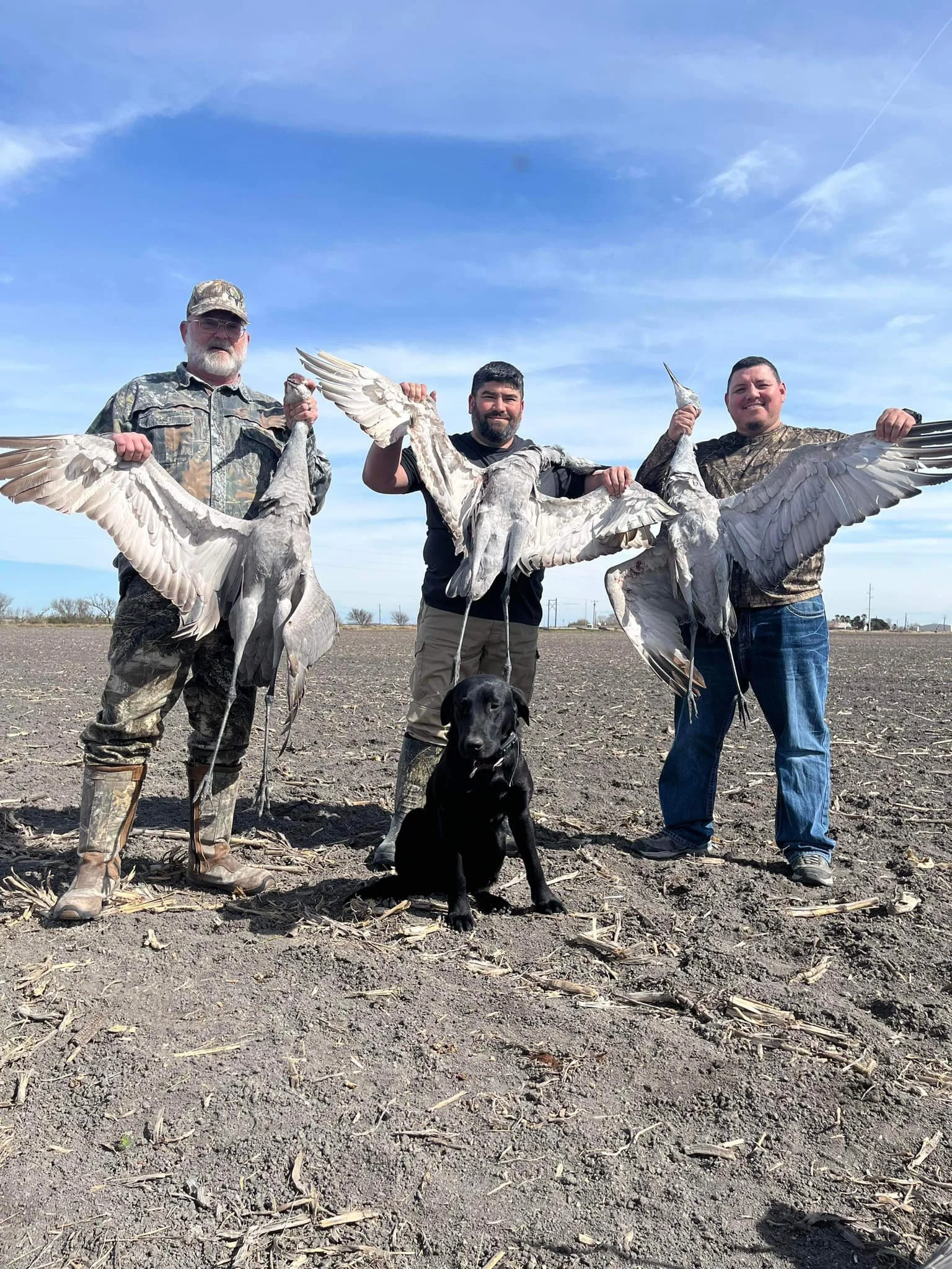 Three hunters holding large birds from a hunting trip in a dirt field, with a black dog sitting in front of them and a clear blue sky overhead.
