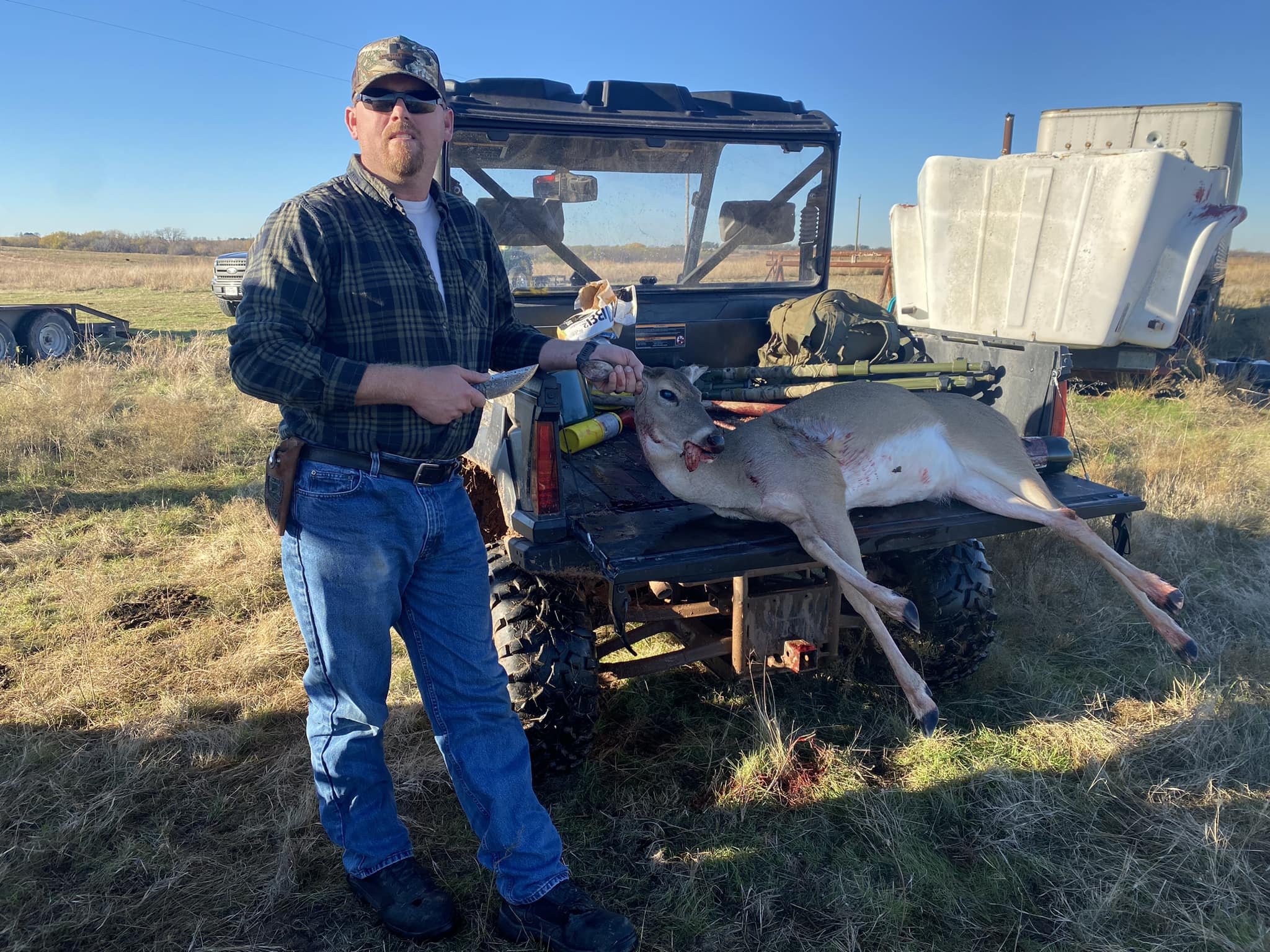 A man in outdoor clothing with sunglasses standing next to a hunting ATV with a freshly hunted deer laid across the back of the vehicle. He is holding a knife and a snack in his hands, and the scene is set in an open field under a clear blue sky.