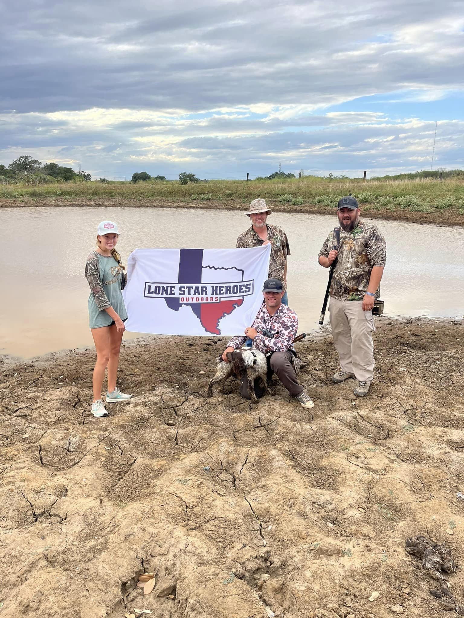 A group of four people, two women and two men, standing outdoors near a muddy pond with grass and trees in the background. They are holding a large flag that reads "Lone Star Heroes Outdoors" with the outline of Texas. One man is kneeling with a dog,