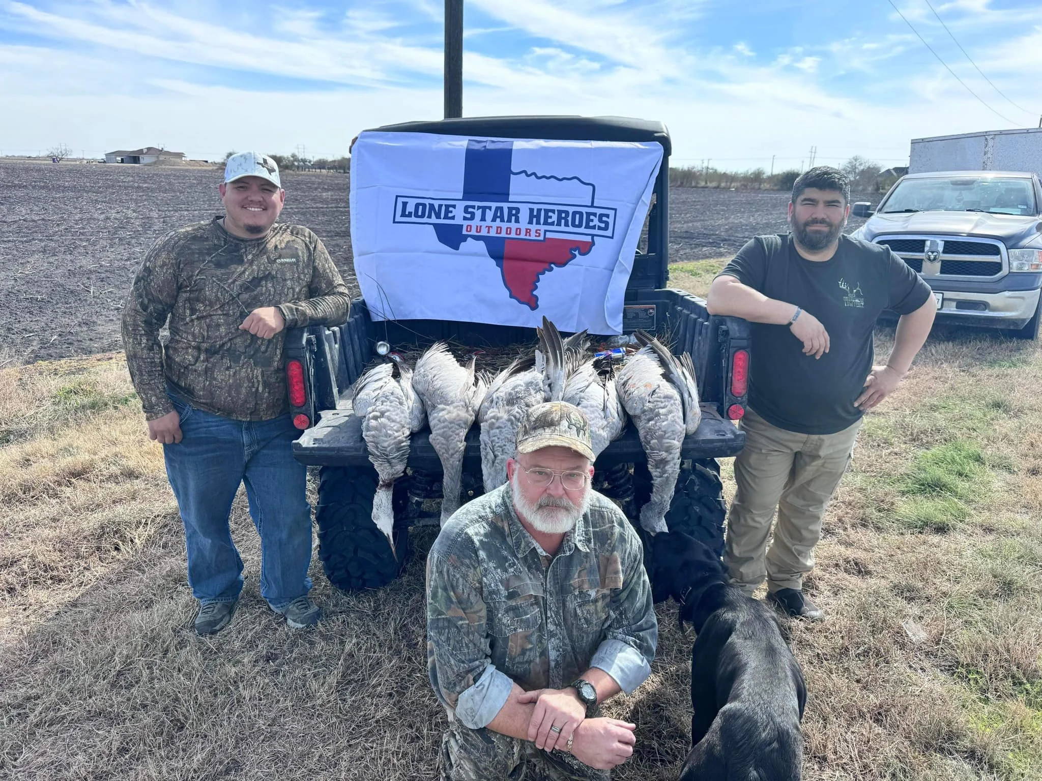Three men with a hunting dog in front of a pickup truck displaying a flag with the Lone Star Heroes Outdoors logo, with several hunted ducks laid on the truck bed, on an open field under a partly cloudy sky.