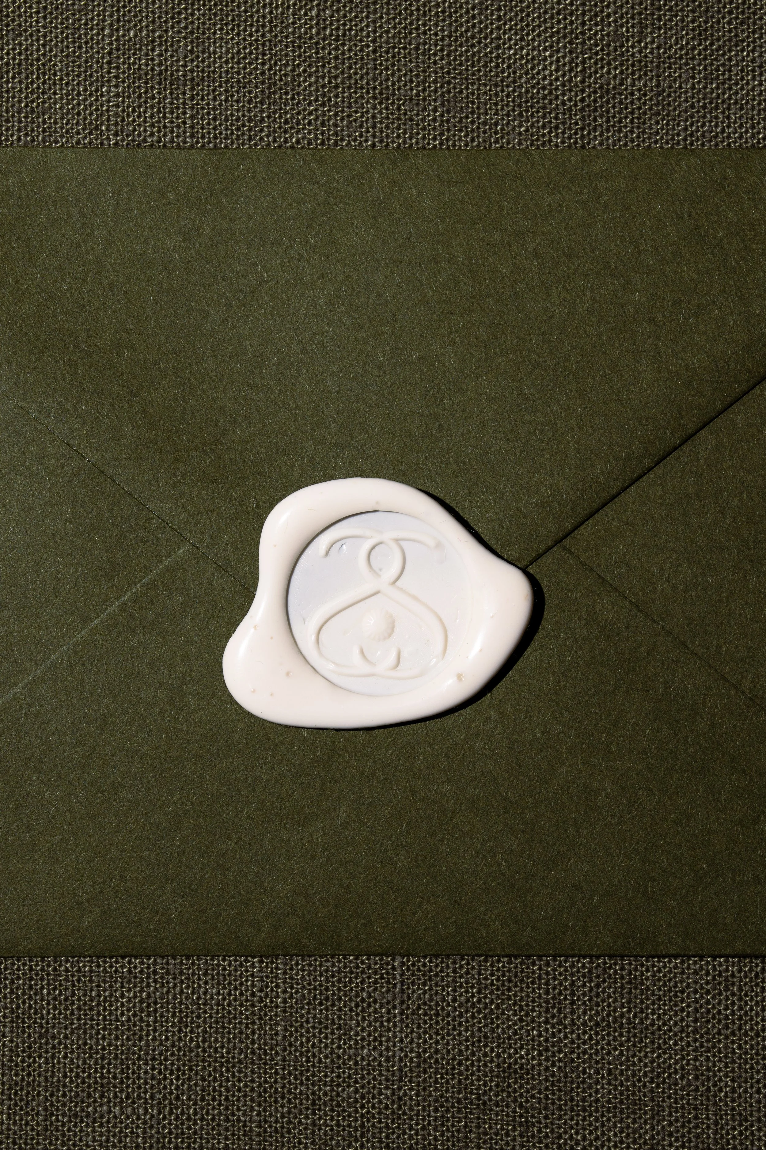 Image of a moss grean linen envelope sealed with a cream wax seal bearing a logo stamp.