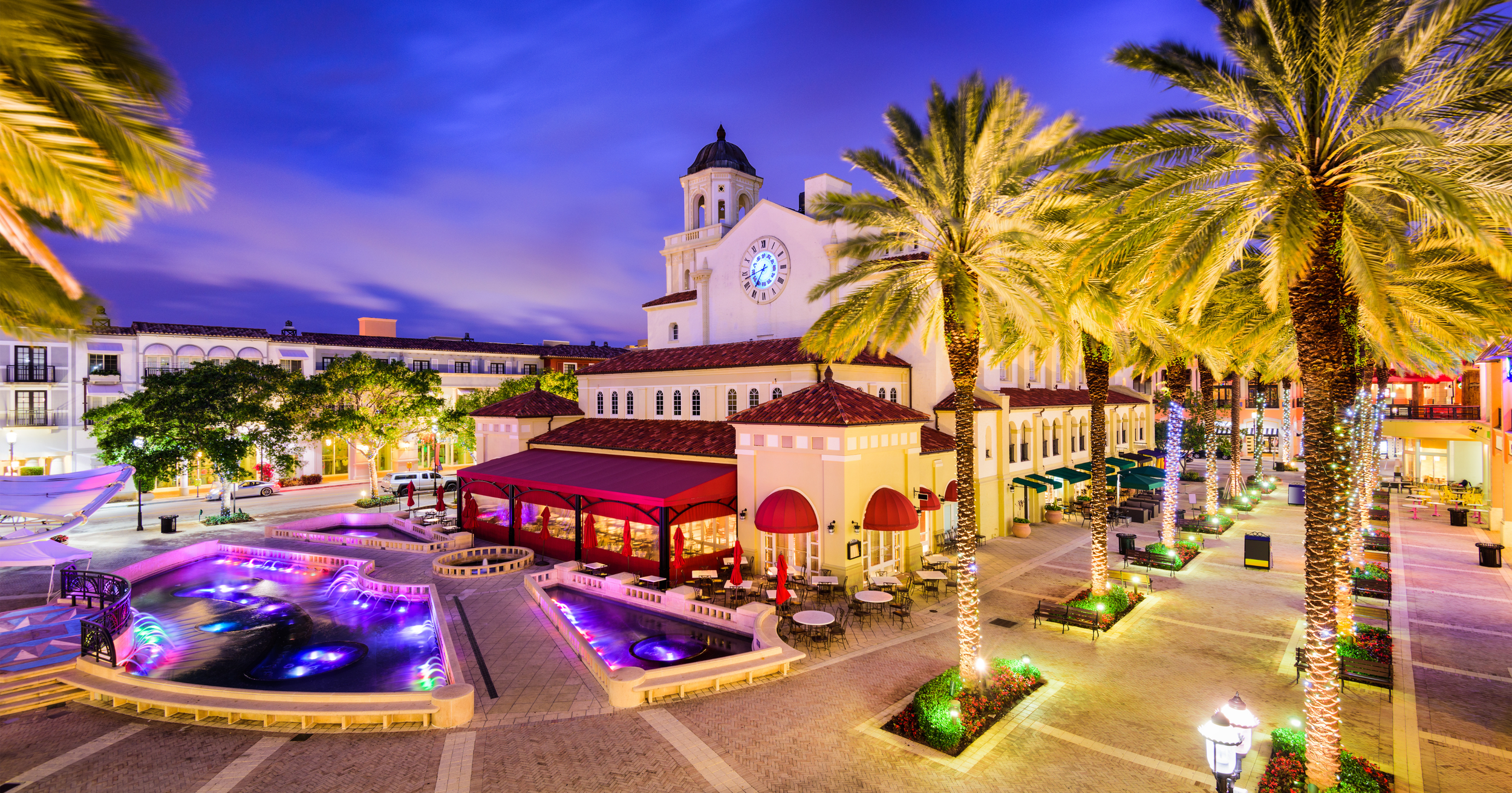 A nighttime view of a lively shopping center with a clock tower, illuminated palm trees, outdoor seating, a water feature, and colorful lights.
