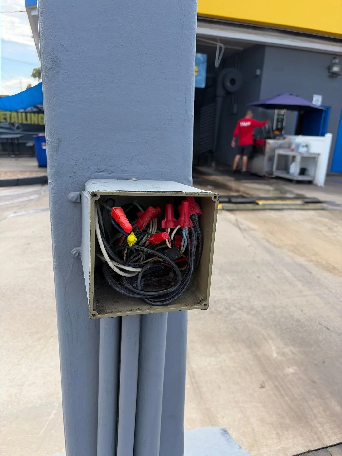 Close-up of an open electrical box with tangled wires, on a grey pole outdoors, with a blurred background showing a person in a red shirt and a yellow and grey building.