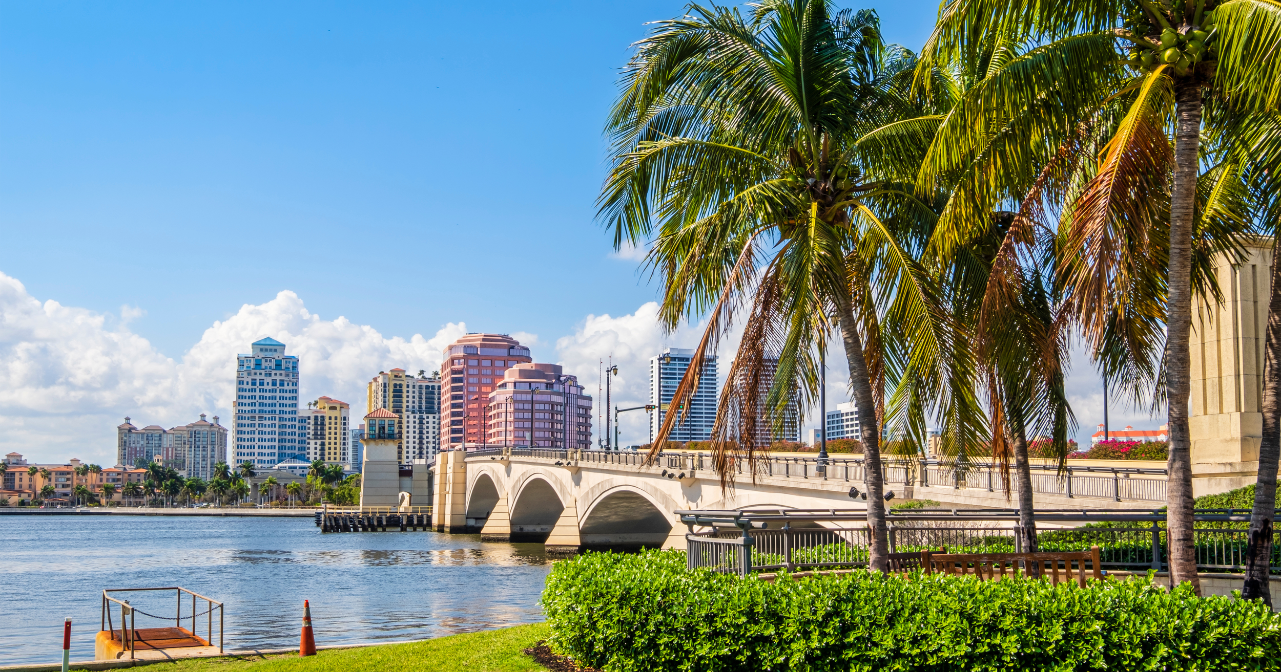 City skyline with colorful high-rise buildings, palm trees in foreground, and a bridge over water on a sunny day with blue sky and clouds.