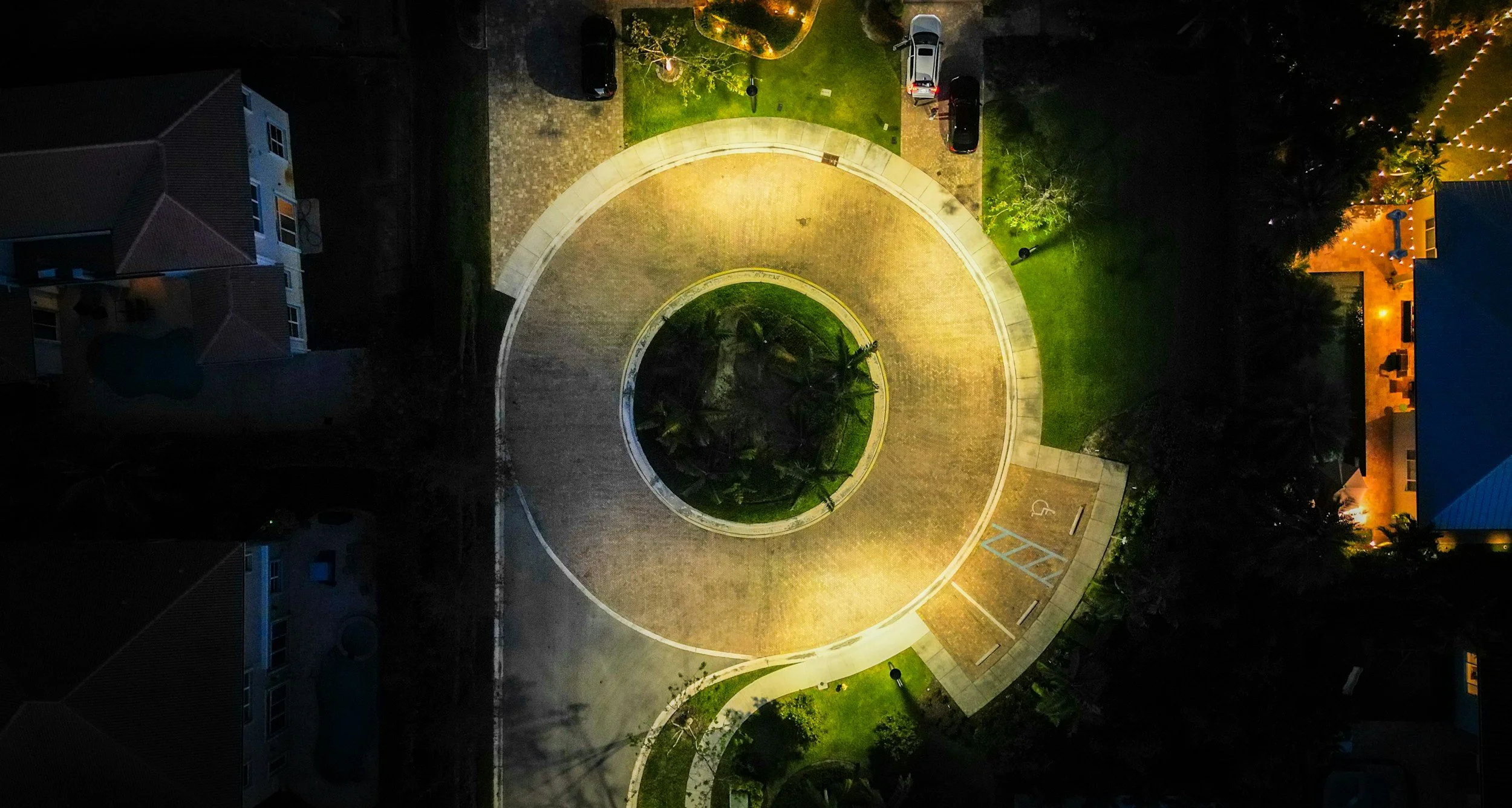 An aerial view of a residential area at night featuring a circular driveway with a central island containing trees, surrounded by houses, parked cars, and landscaping, illuminated by streetlights and exterior lighting.
