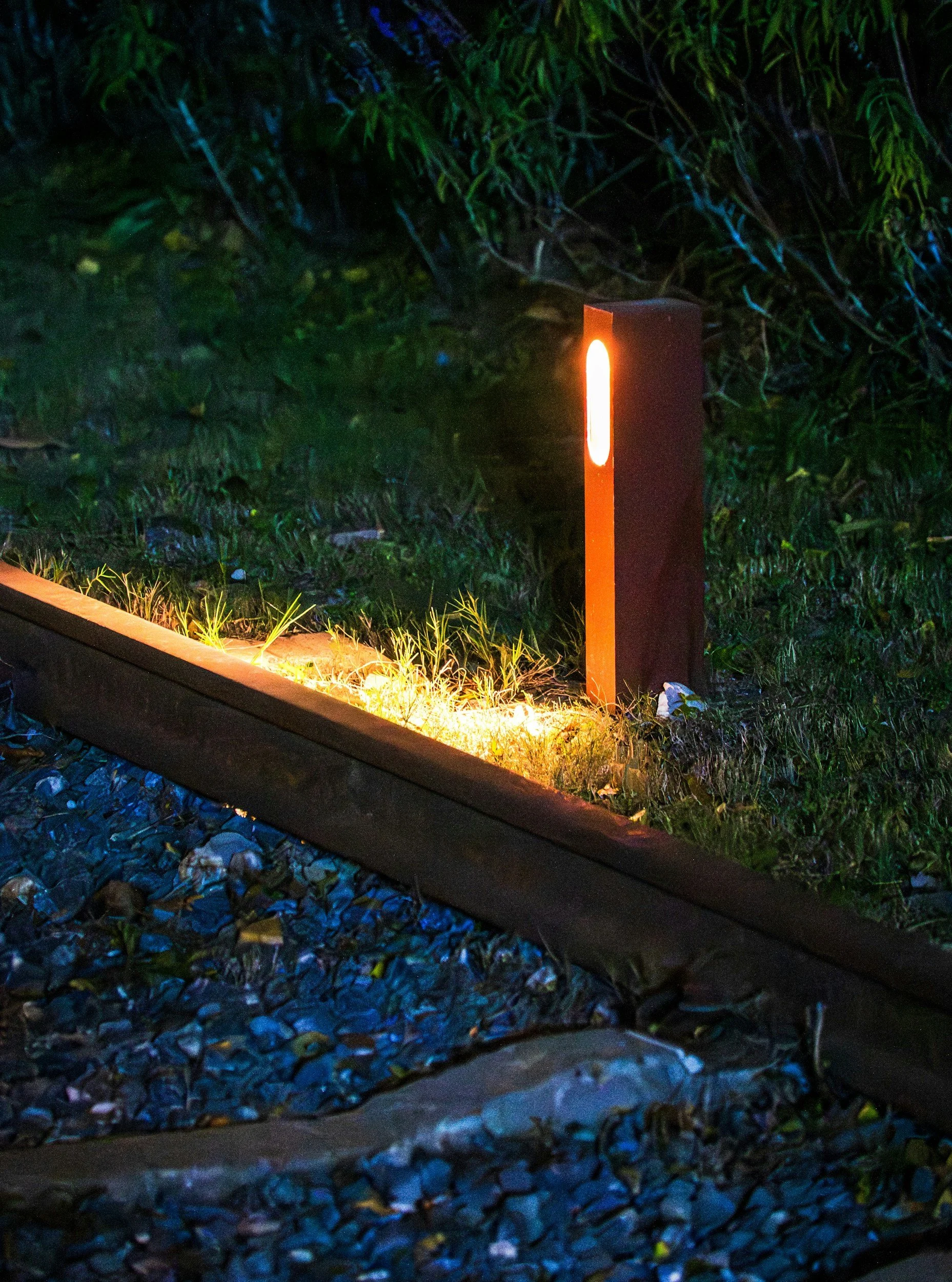 A glowing orange-lit bollard light next to railway tracks and green plants at night.